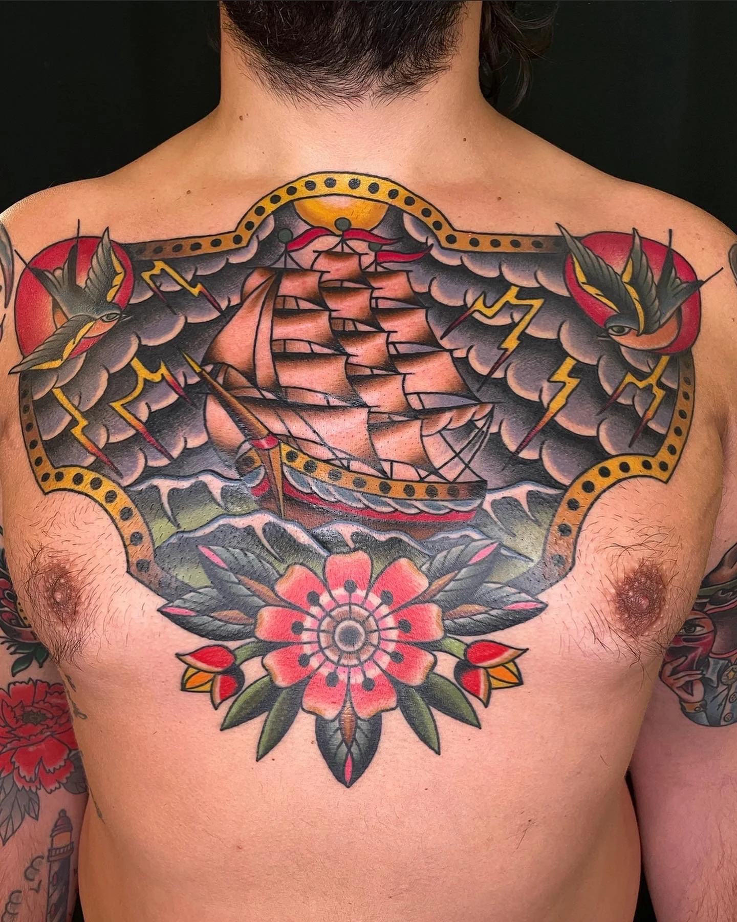 Colorful tattoo of a sailing ship on stormy seas, flanked by two swallows, with a flower design below the ship, on a person's chest.