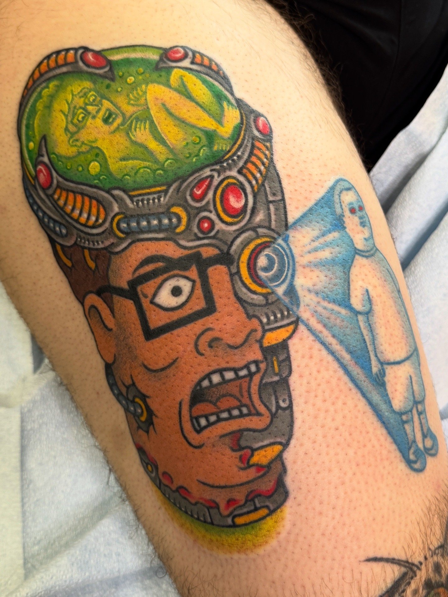 A colorful tattoo featuring a woman with glasses and earrings, with an open-mouthed expression, wearing a high-tech helmet and breathing apparatus. A small figure with a halo appears from the helmet's other side.