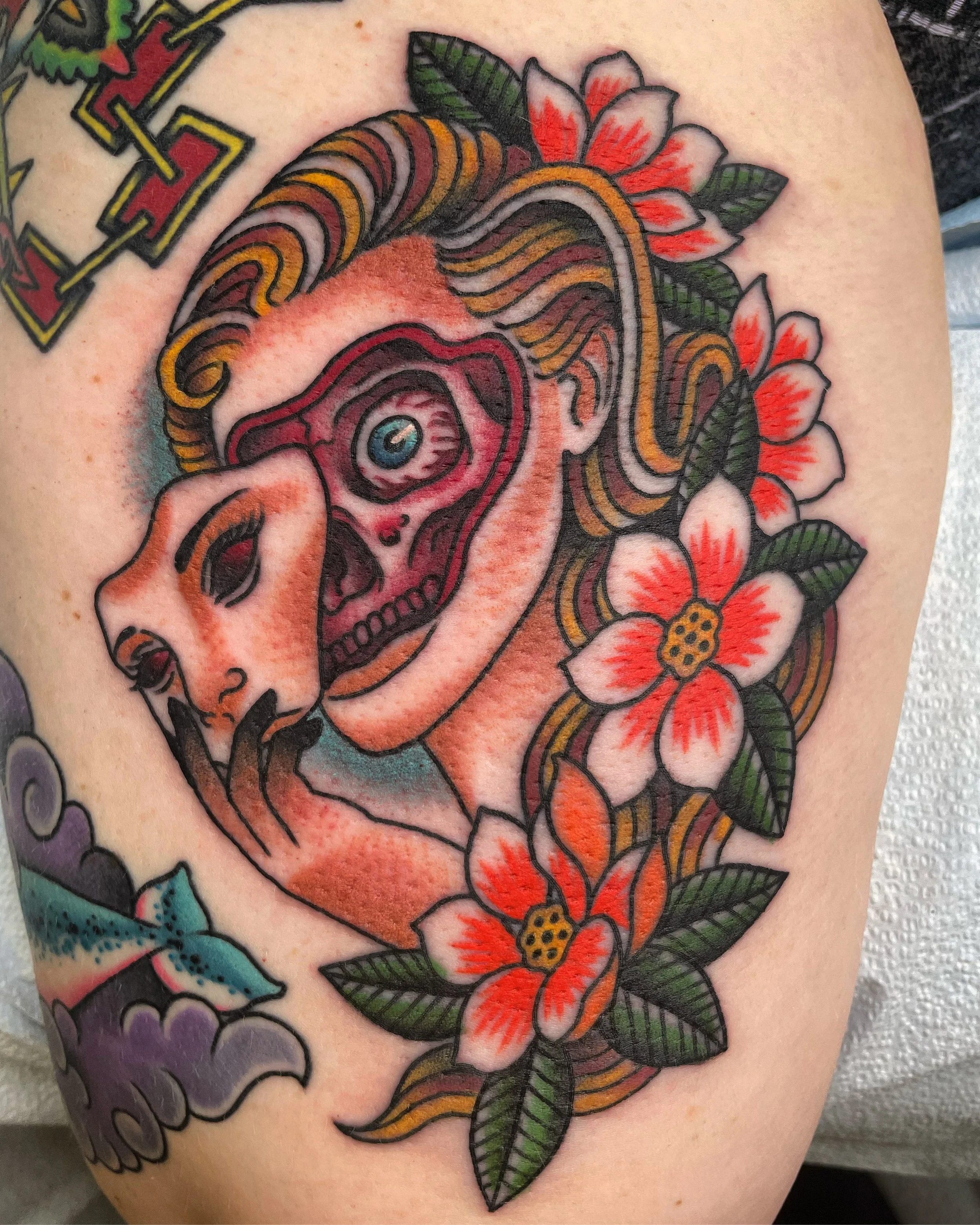 Tattoo of a woman with long wavy hair surrounded by pink and orange flowers, with part of a colorful geometric design visible in the top left corner.