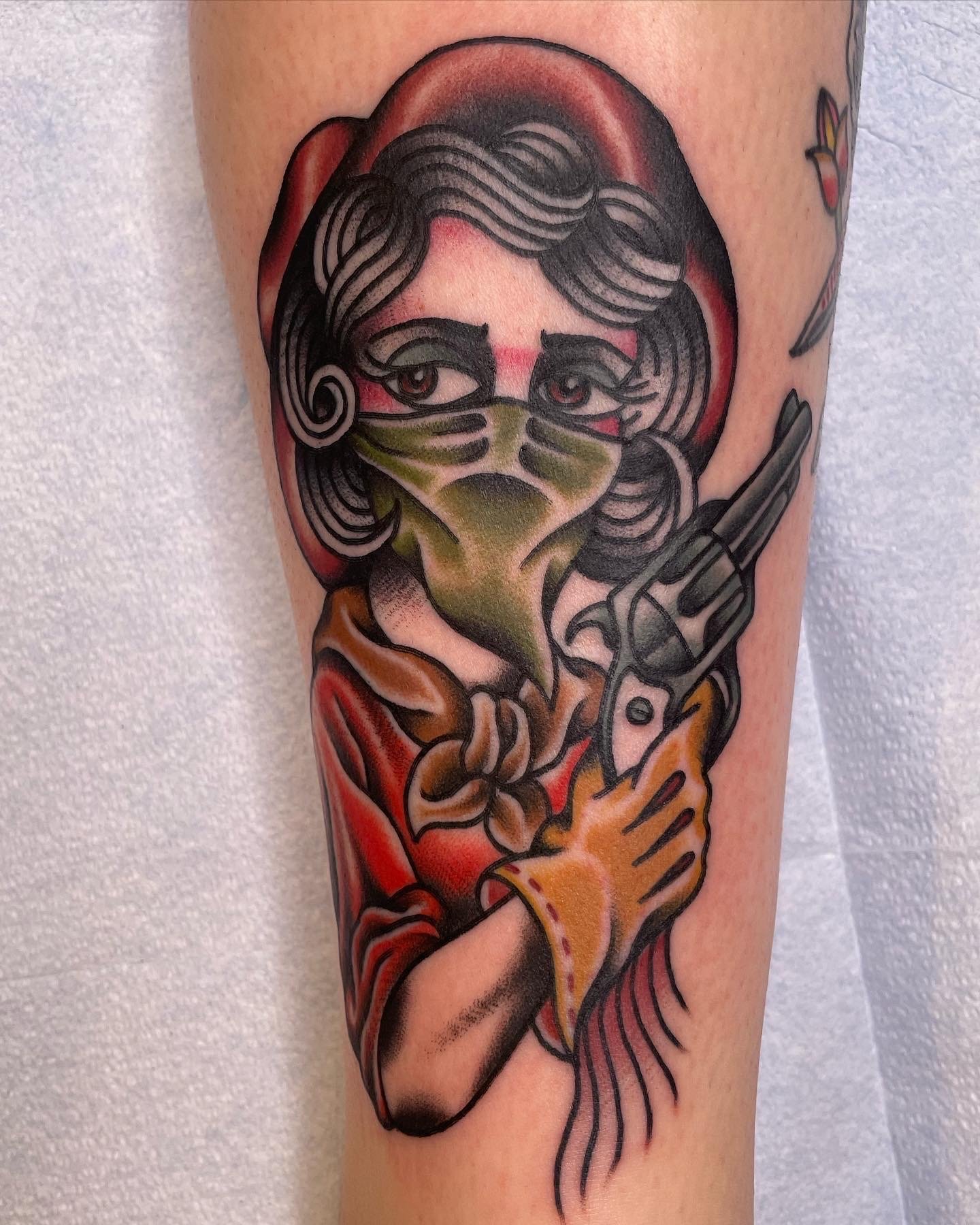 Tattoo of a woman with curly hair and a face mask, holding a revolver, on a person's arm.