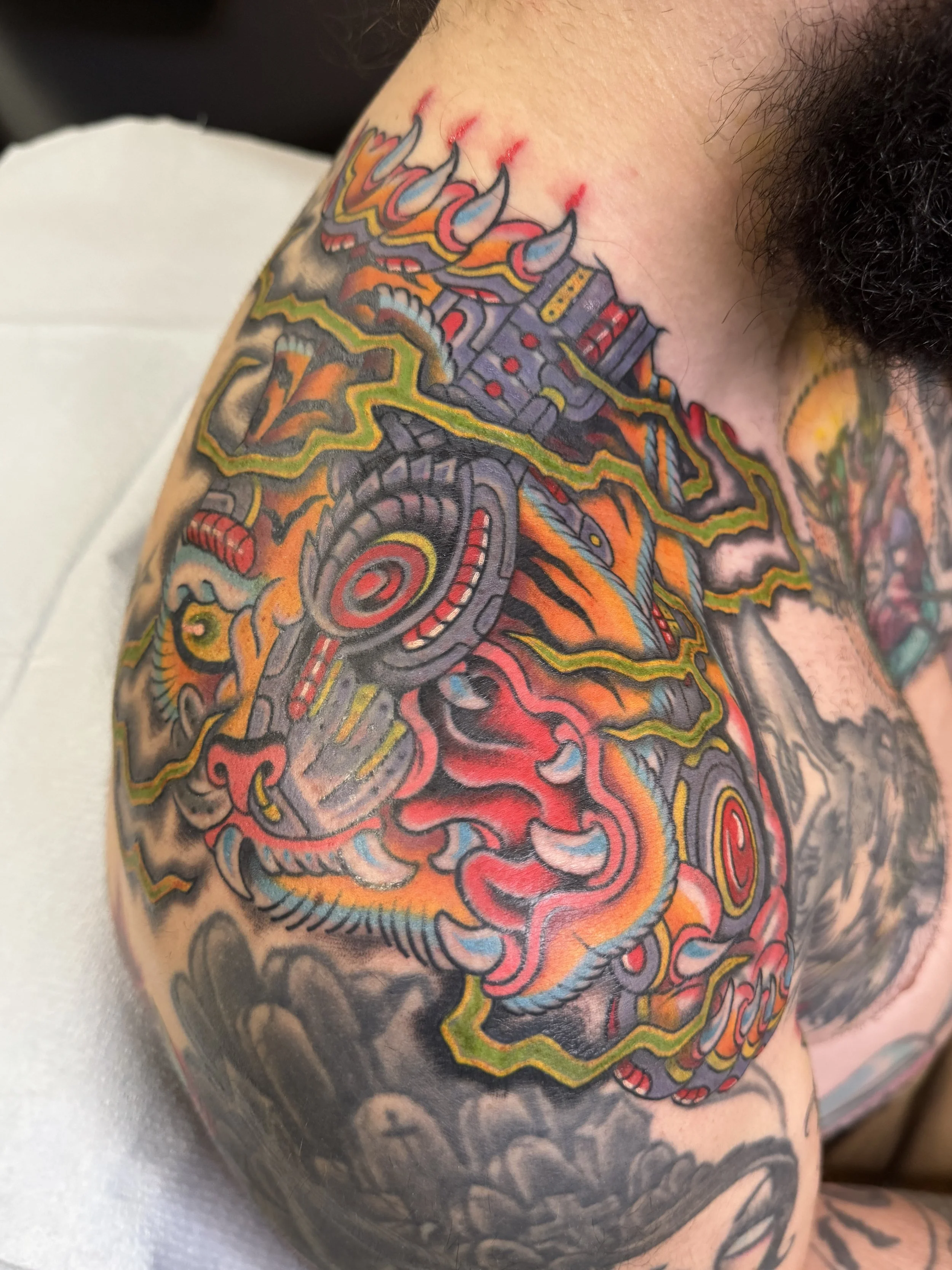 Close-up of a colorful tattoo on a person's shoulder and upper arm featuring a dragon, mechanical elements, and abstract flames in red, orange, yellow, black, gray, and blue colors.