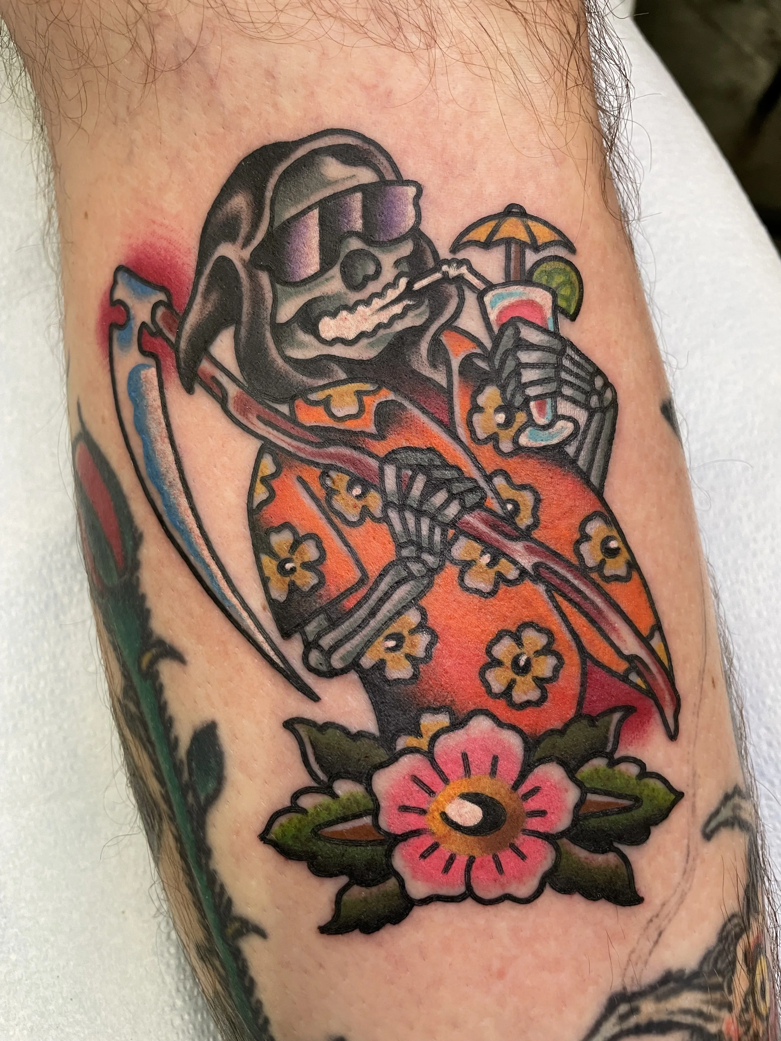 A tattoo of a skeleton wearing sunglasses and a floral-patterned Hawaiian shirt, holding a drink with a tiny umbrella and lime, while sitting with a surfboard, surrounded by colorful flowers.