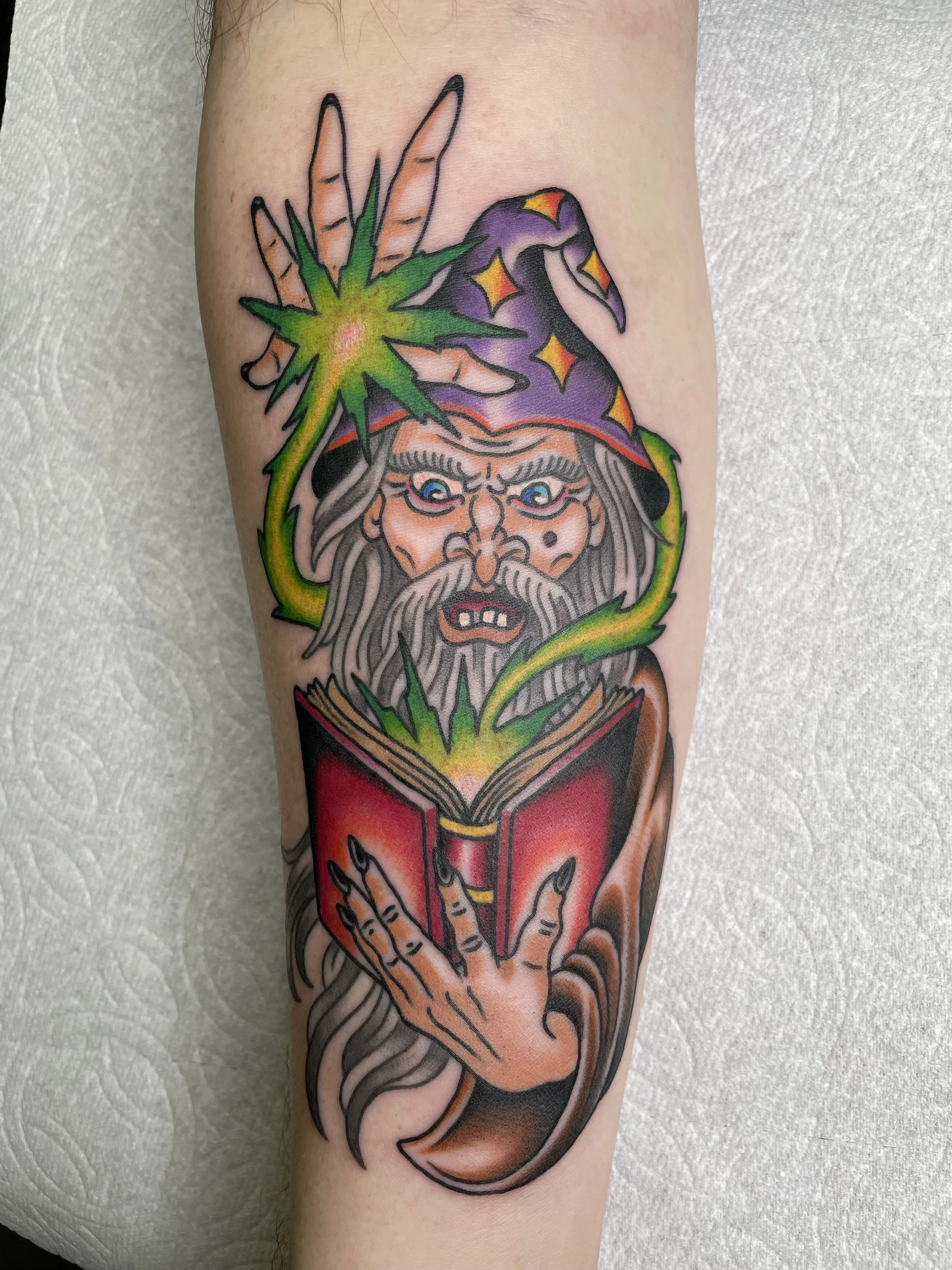 A colorful tattoo of a wizard with a long beard, wearing a starry purple hat, holding a glowing red book, with green magical energy coming from his hand and the book.
