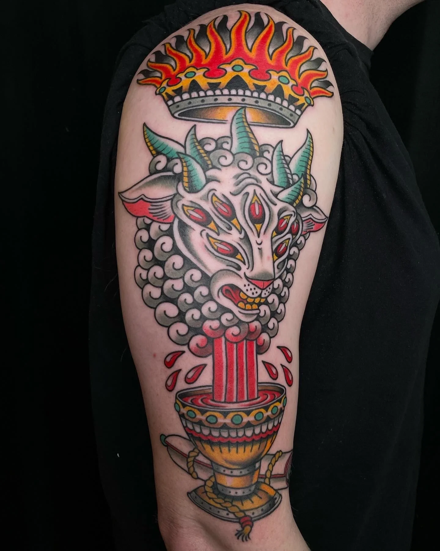 A colorful tattoo of a goat's head with multiple eyes, horns, and flames emerging from the top, blended with a traditional-style chalice that appears to be overflowing and releasing a cloud of smoke or steam.