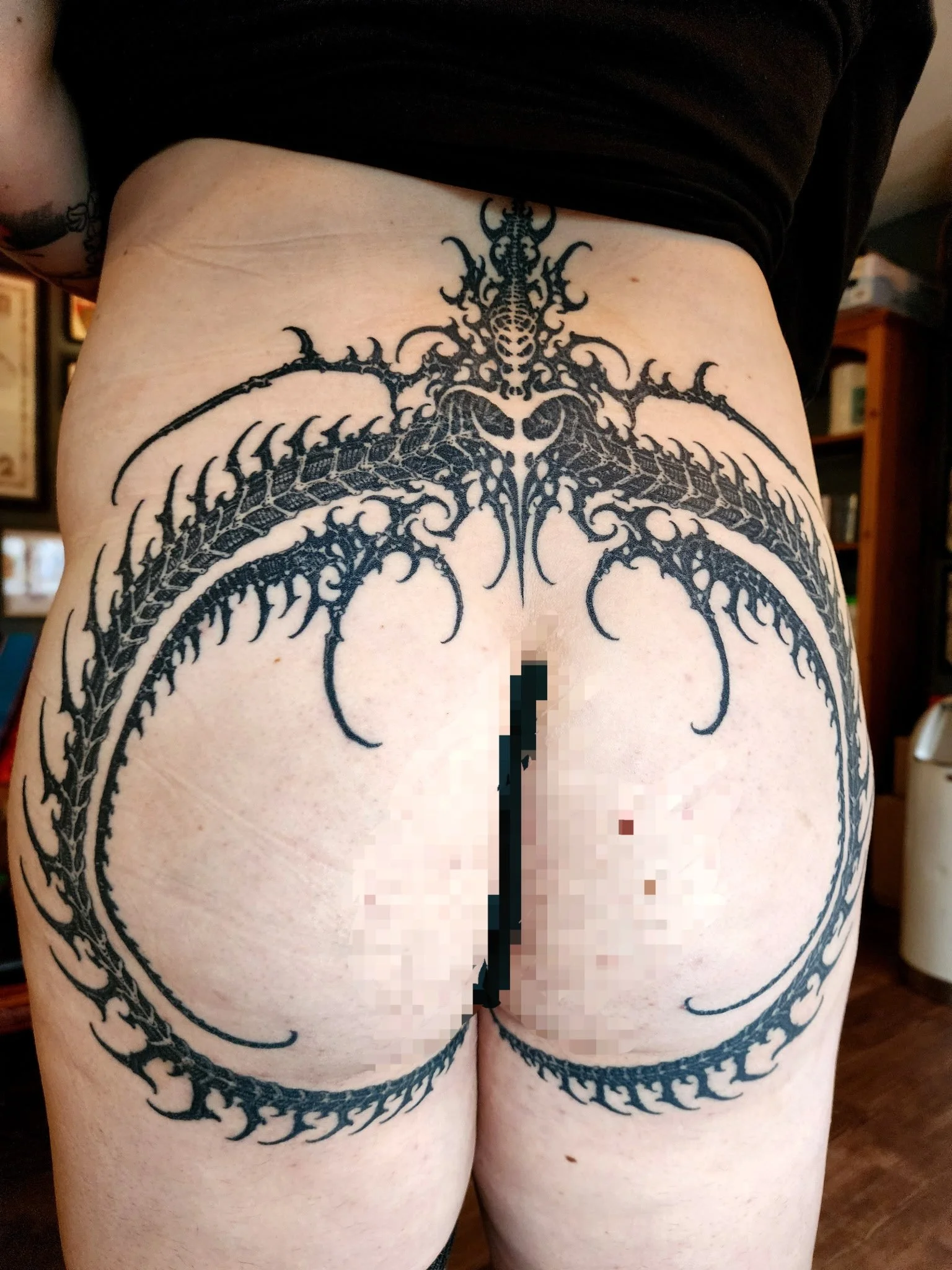 A person's lower back and buttocks with a large black tattoo of a monstrous, symmetrical dragon-like creature with angular features, wings, and multiple intricate details.
