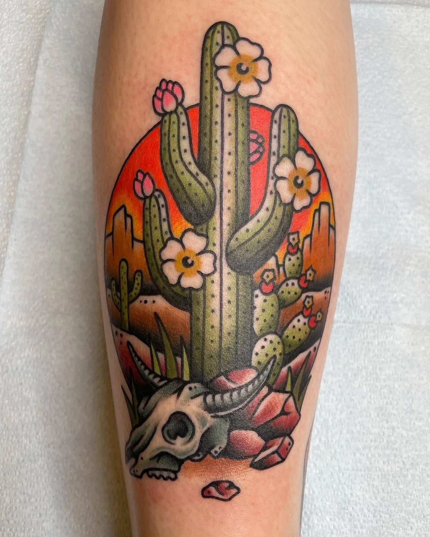 Tattoo of a desert scene with a large cactus, desert flowers, mountains, and a cow skull on rocks.