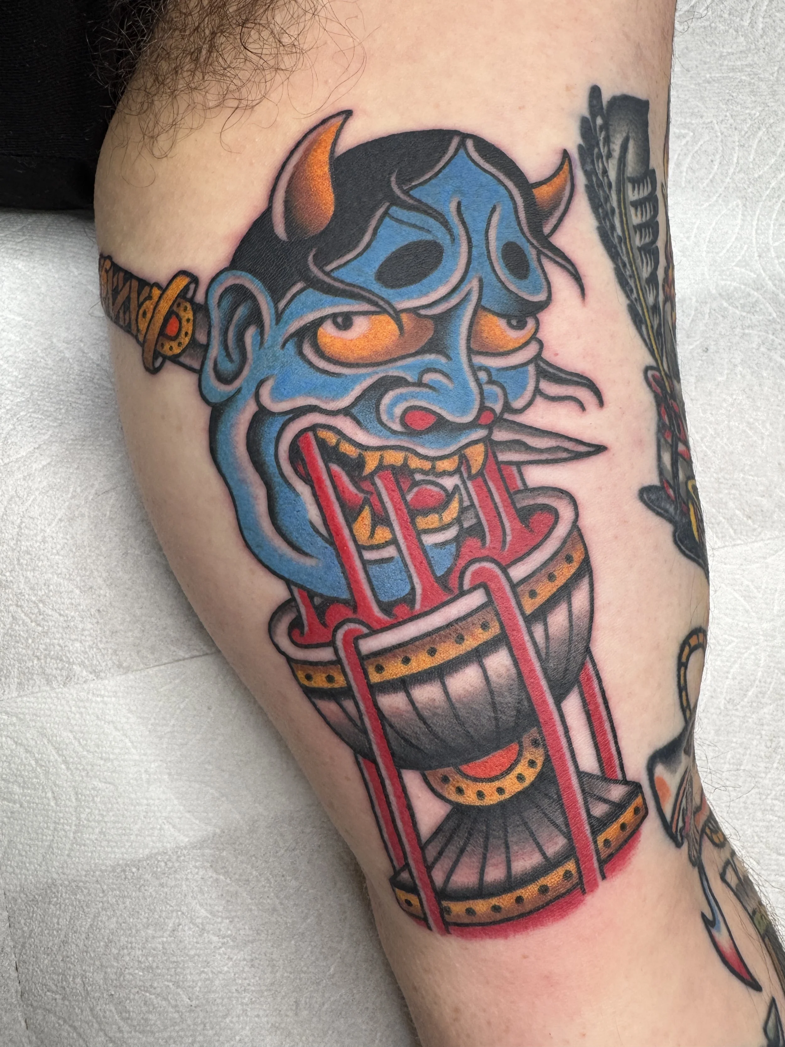 Tattoo of a blue demon face with orange horns, dripping red liquid from its eyes and mouth, above a traditional-style drum.