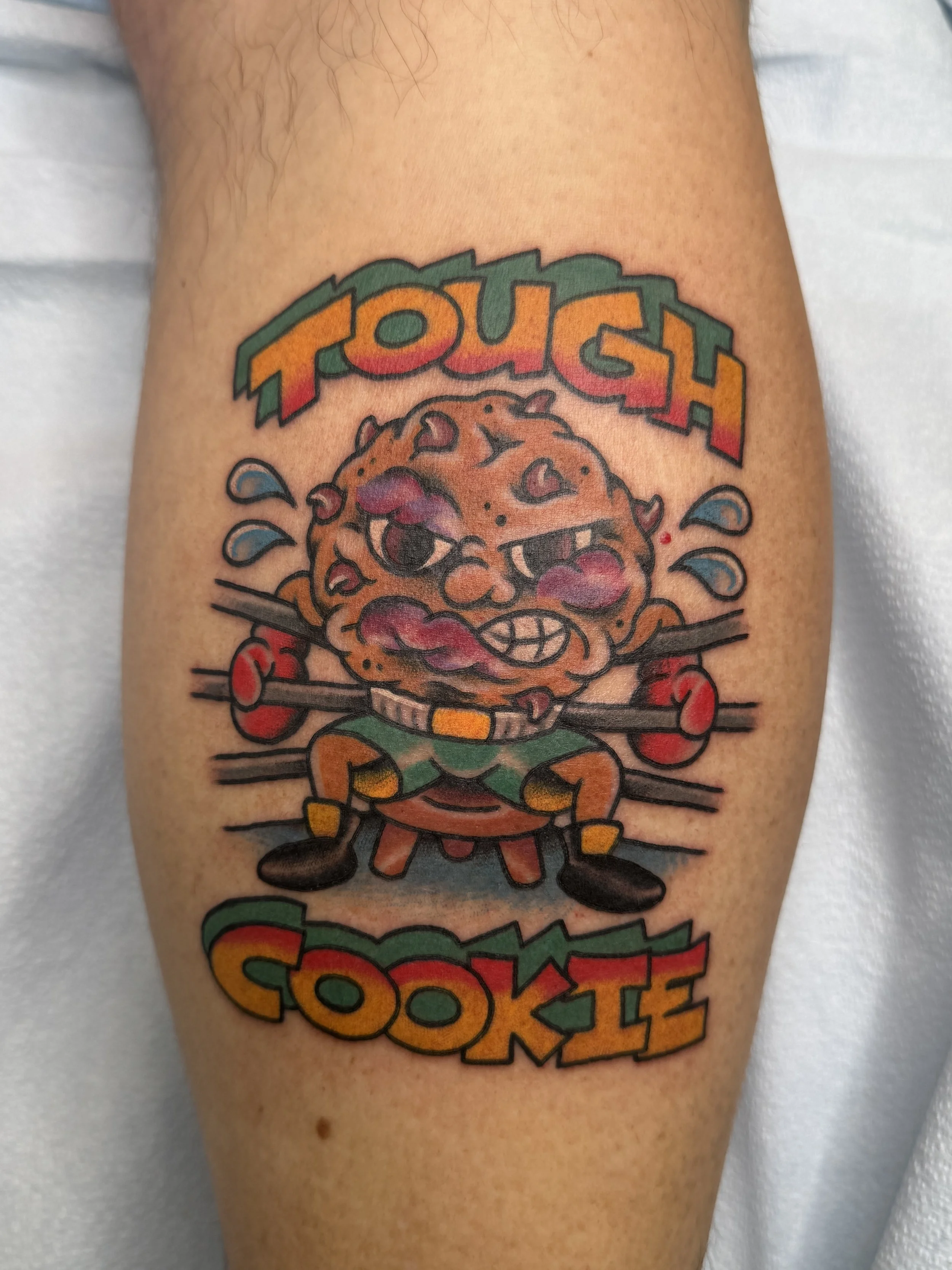 Colorful tattoo of a cartoon cookie character with boxing gloves and shorts, sweating, with the text 'TOUGH COOKIE' above and below.