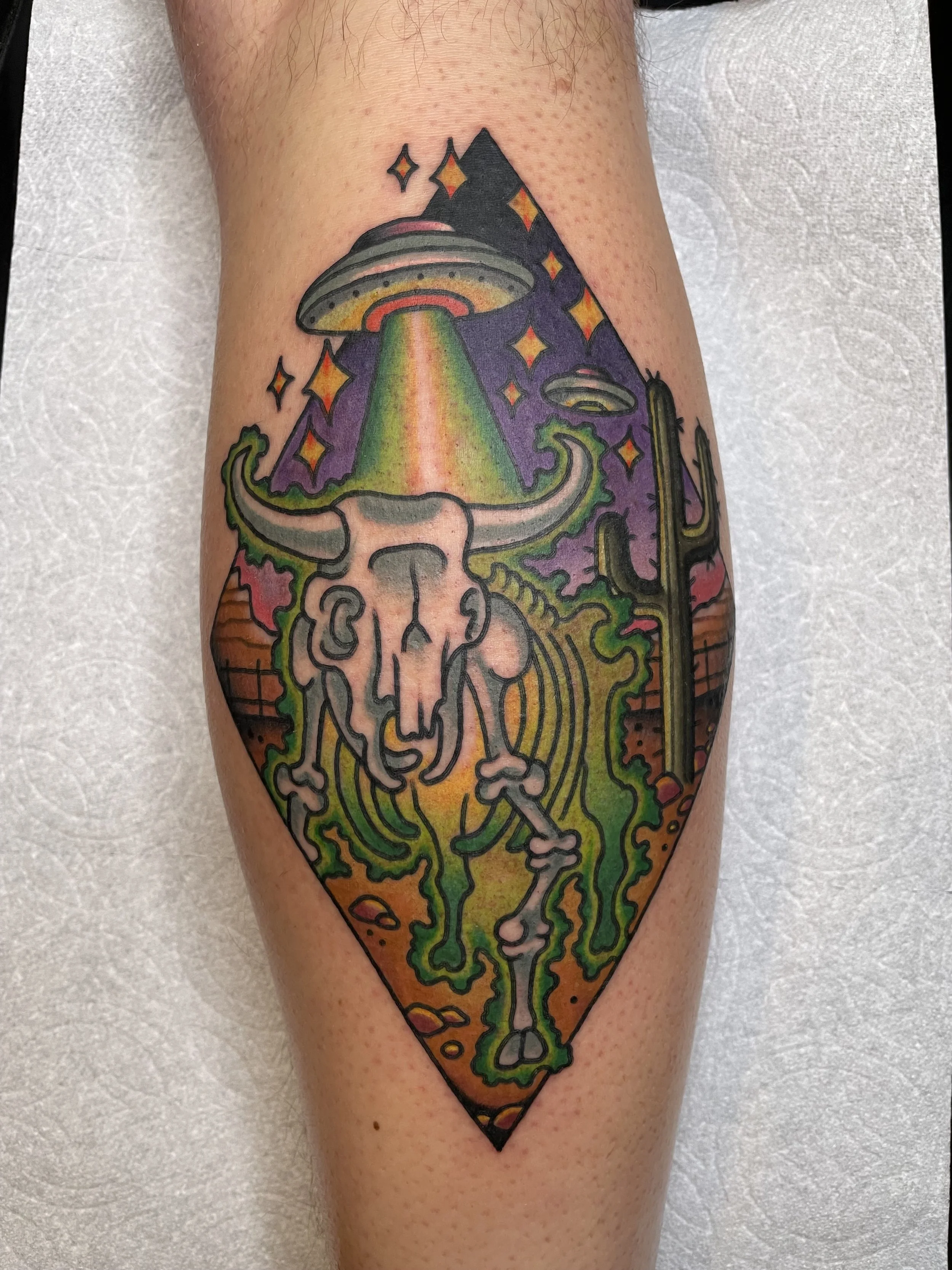 Colorful tattoo of a bison skull with horns, topped by a rainbow-colored UFO beam, surrounded by an alien landscape with cactus, mountains, and an alien spaceship in the sky, all within a diamond-shaped frame.