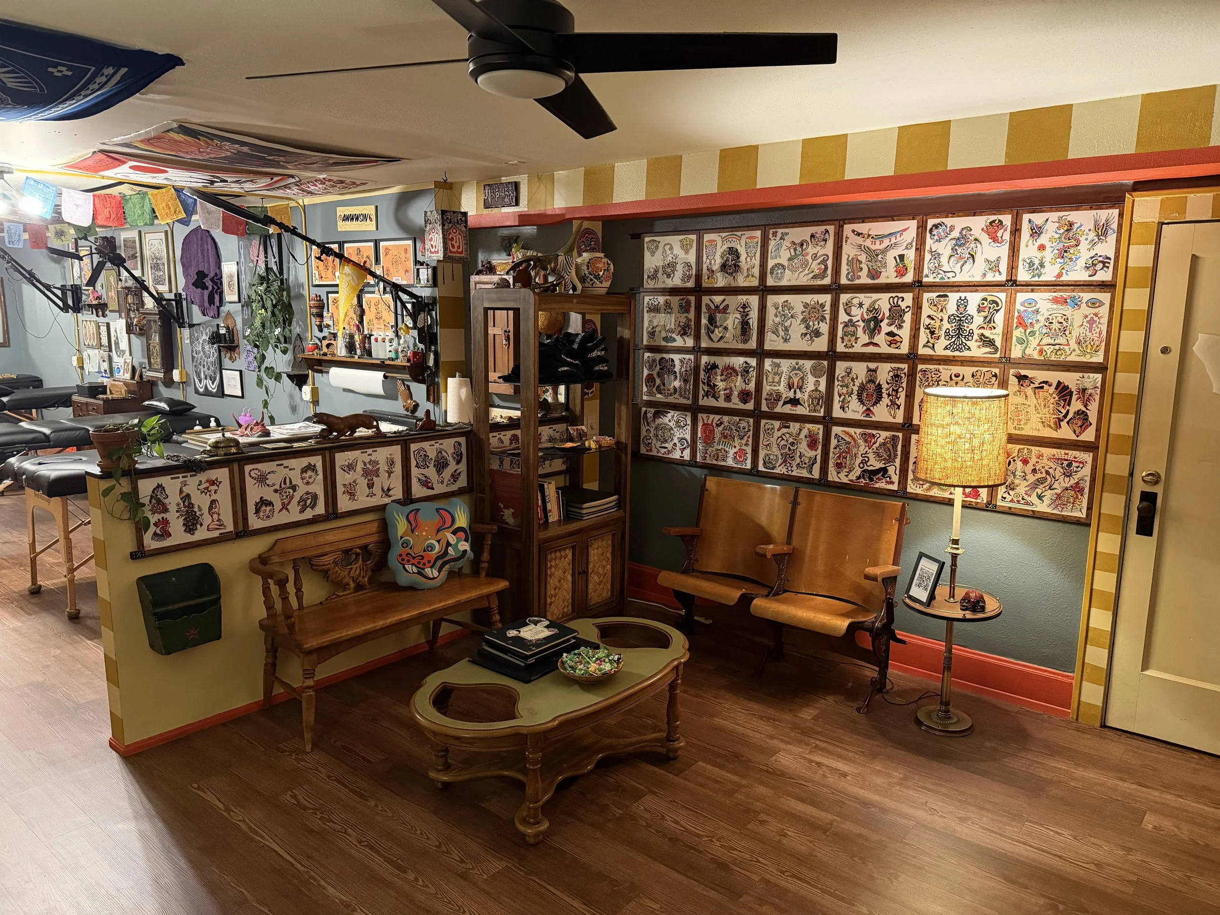 Cozy tattoo studio corner with framed tattoo art on the wall, vintage wooden furniture, a floor lamp, and tattoo equipment.