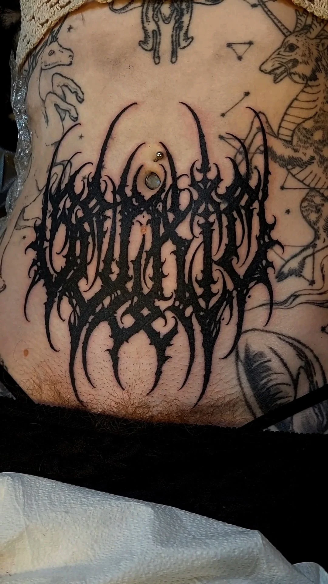 Close-up of a person's chest with a black tattoo of an intricate, spiky, band logo style design, surrounded by other tattoos including a lion, a snake, a goat, and a wolf, as well as piercings.
