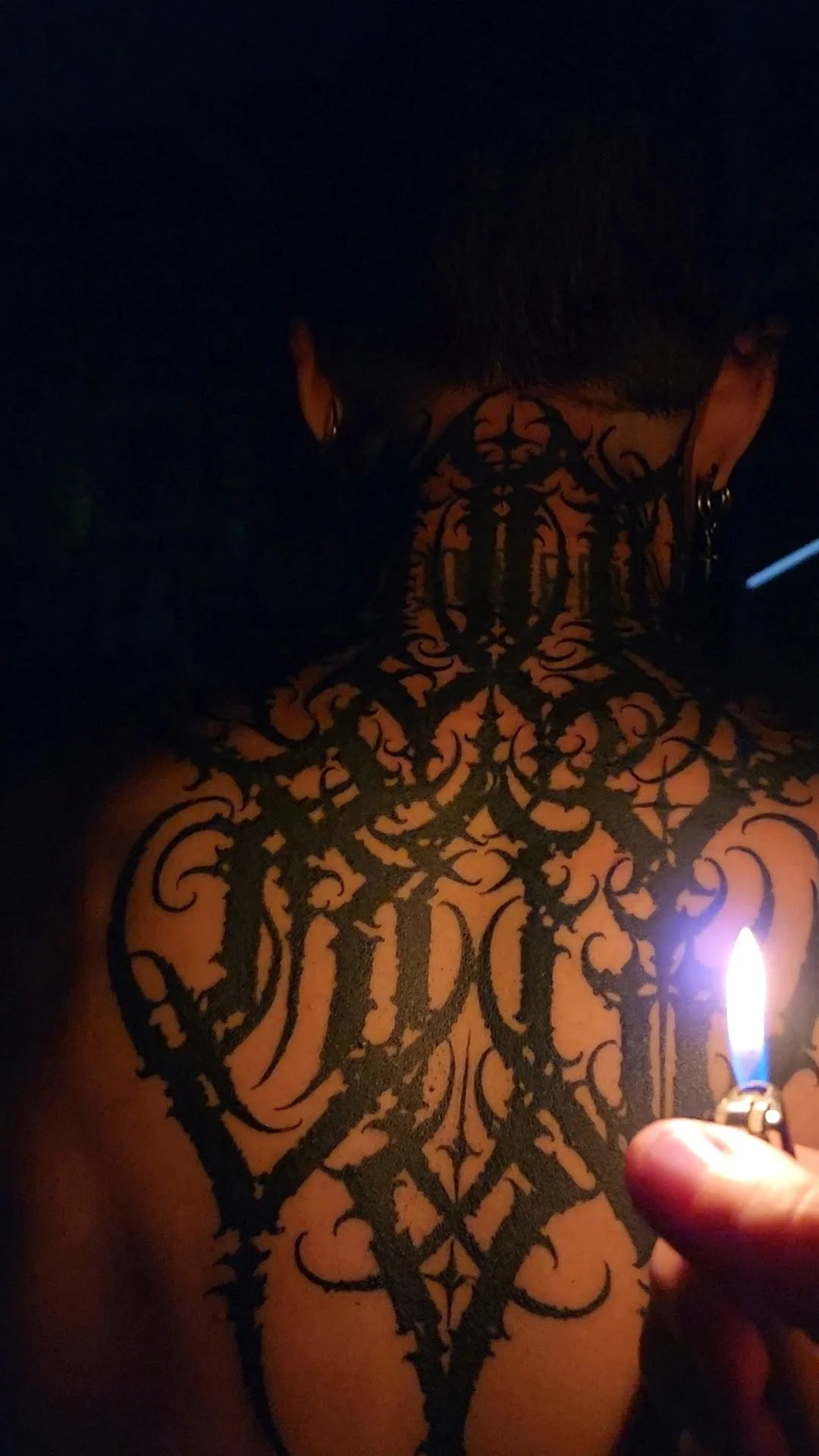 Person with extensive black tattoos covering their neck and upper back, holding a lit lighter in a dark environment.