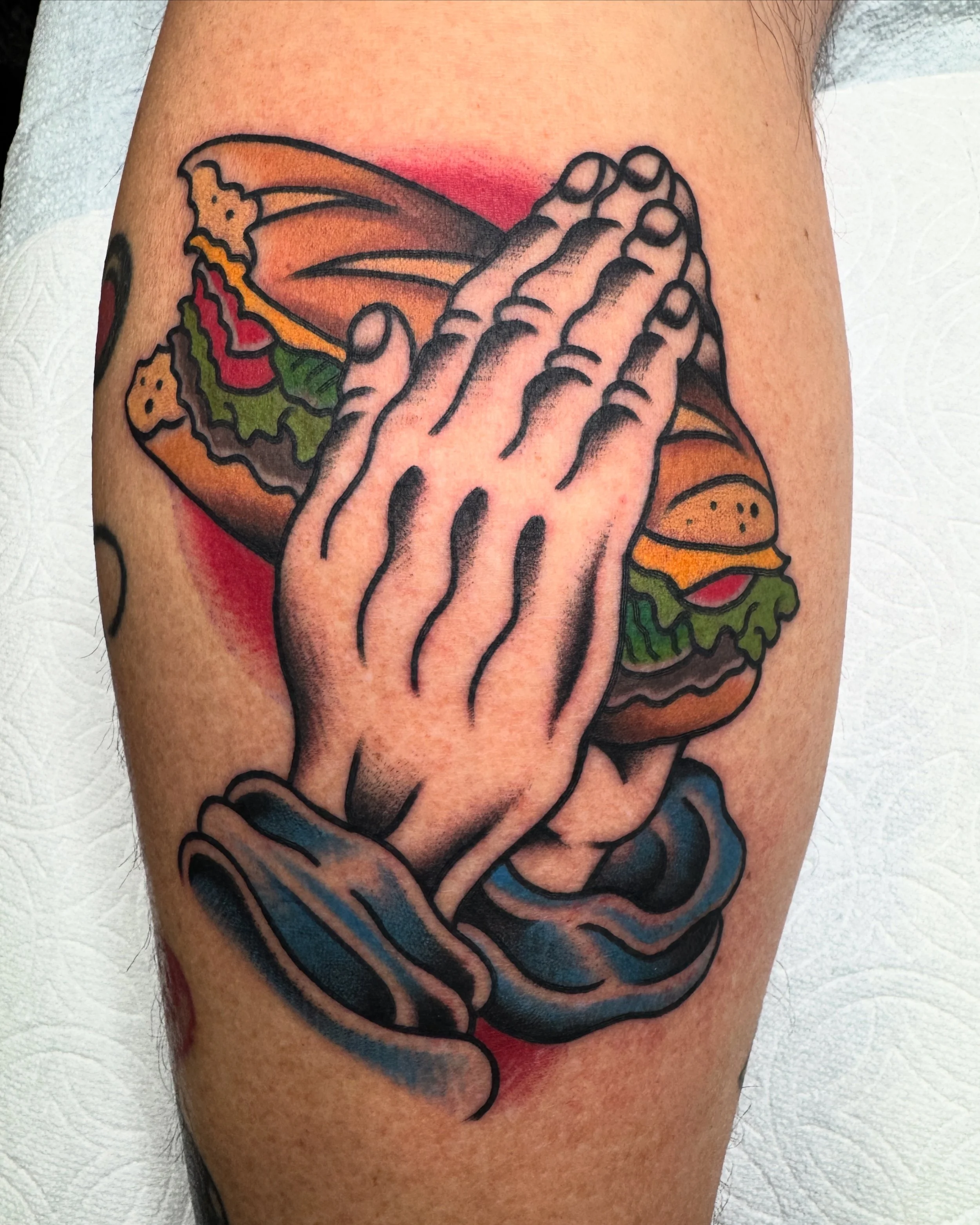 Tattoo of praying hands holding a hamburger with lettuce, tomato, and cheese.