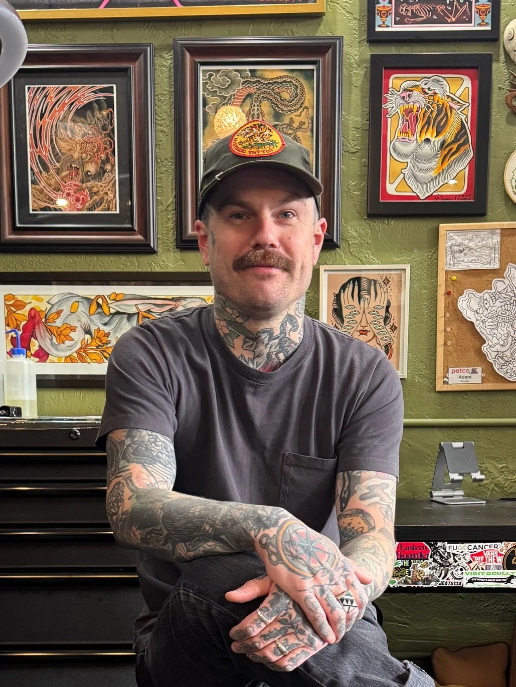 A man with tattoos on his arms, neck, and hands sitting in front of a green wall decorated with various framed tattoo art and illustrations, wearing a dark t-shirt and a hat with a tiger patch.