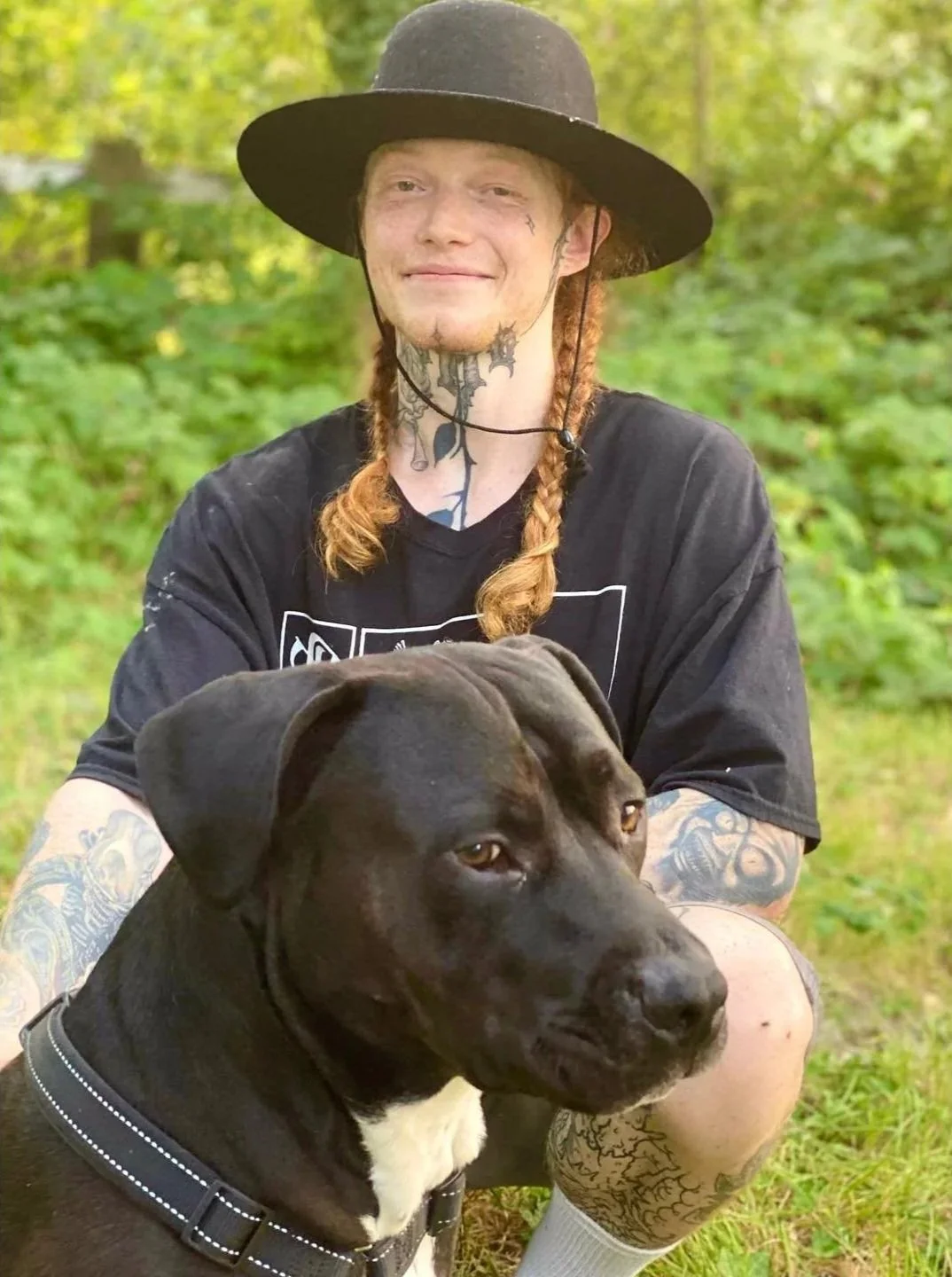 A young man with tattoos, long hair in braids, and a wide-brimmed black hat, sitting outdoors with a large black and white dog.