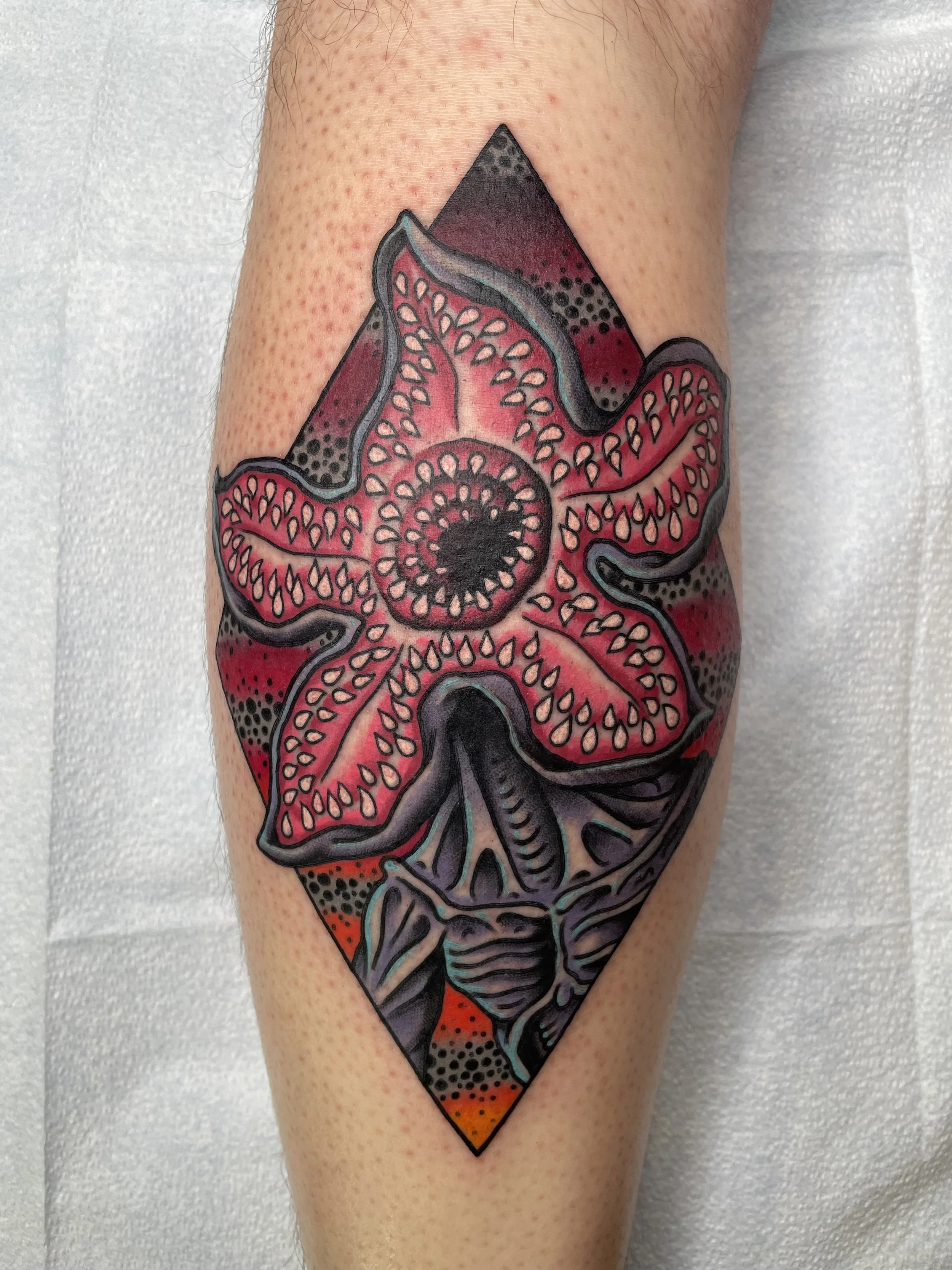 A colorful tattoo of a pink and red flower with black and blue outlines on a person's arm, set within a diamond-shaped background with dotted shading.