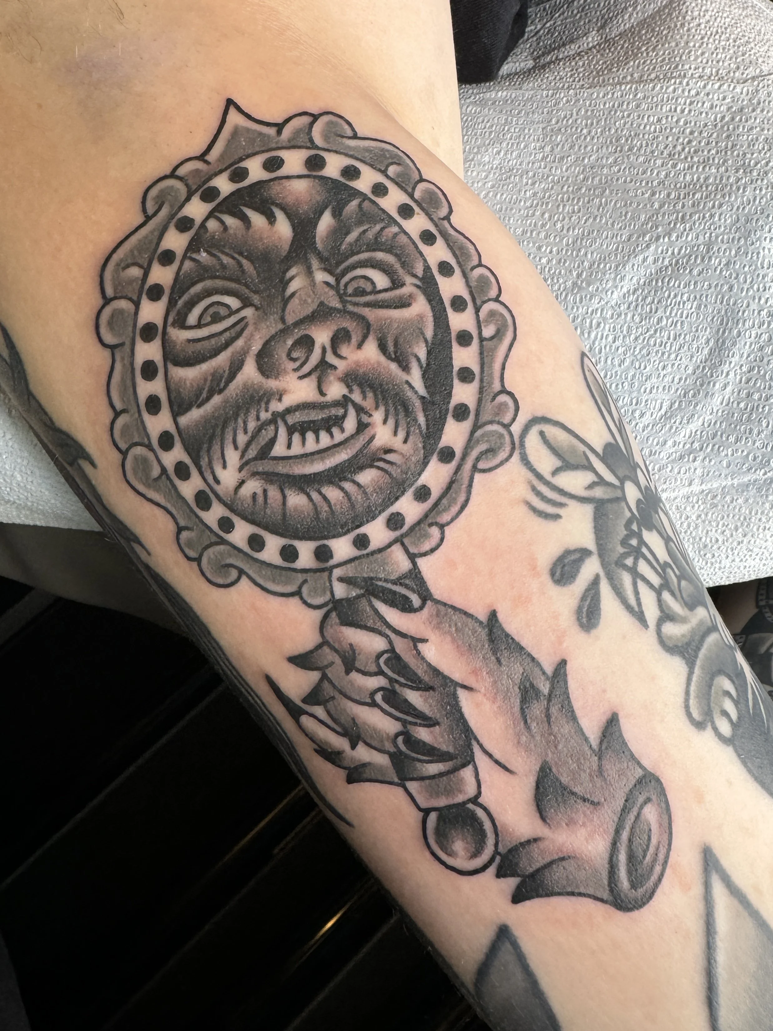 Tattoo of a snarling face with a mirror-like frame and chain link on the arm.
