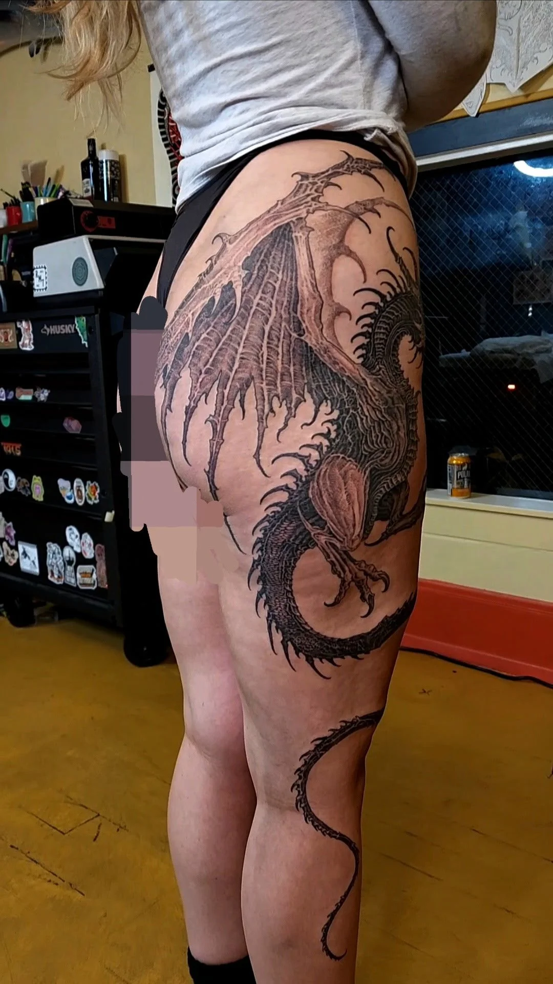 A woman with a large, detailed black tattoo of a dragon on her right thigh and hip, extending onto her lower stomach.