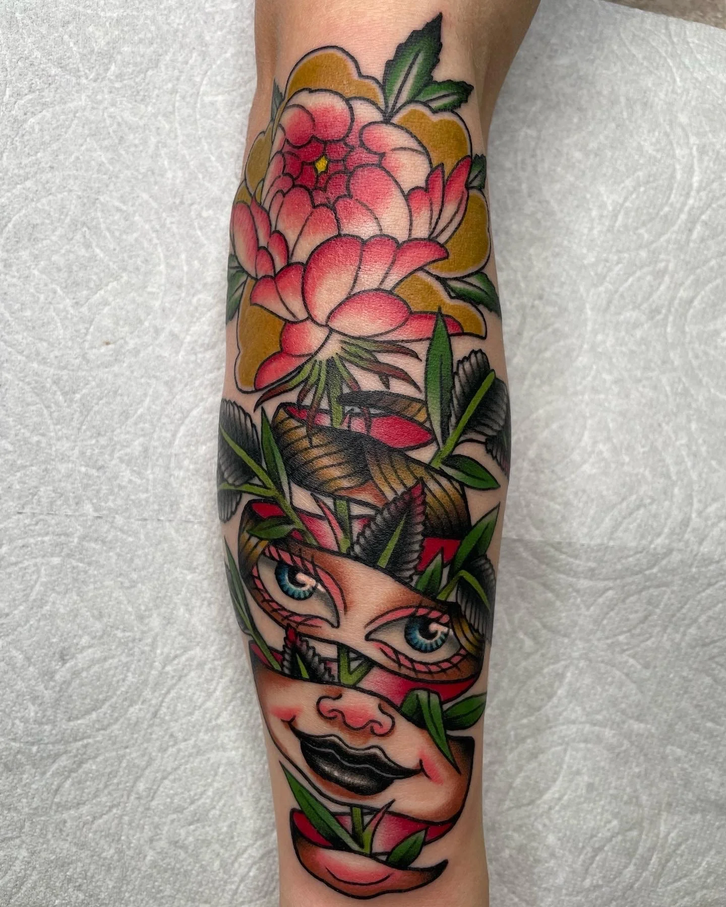 Colorful tattoo of a woman's face with blue eyes, black lips, and a fox mask, surrounded by pink flowers, green leaves, and black accents.