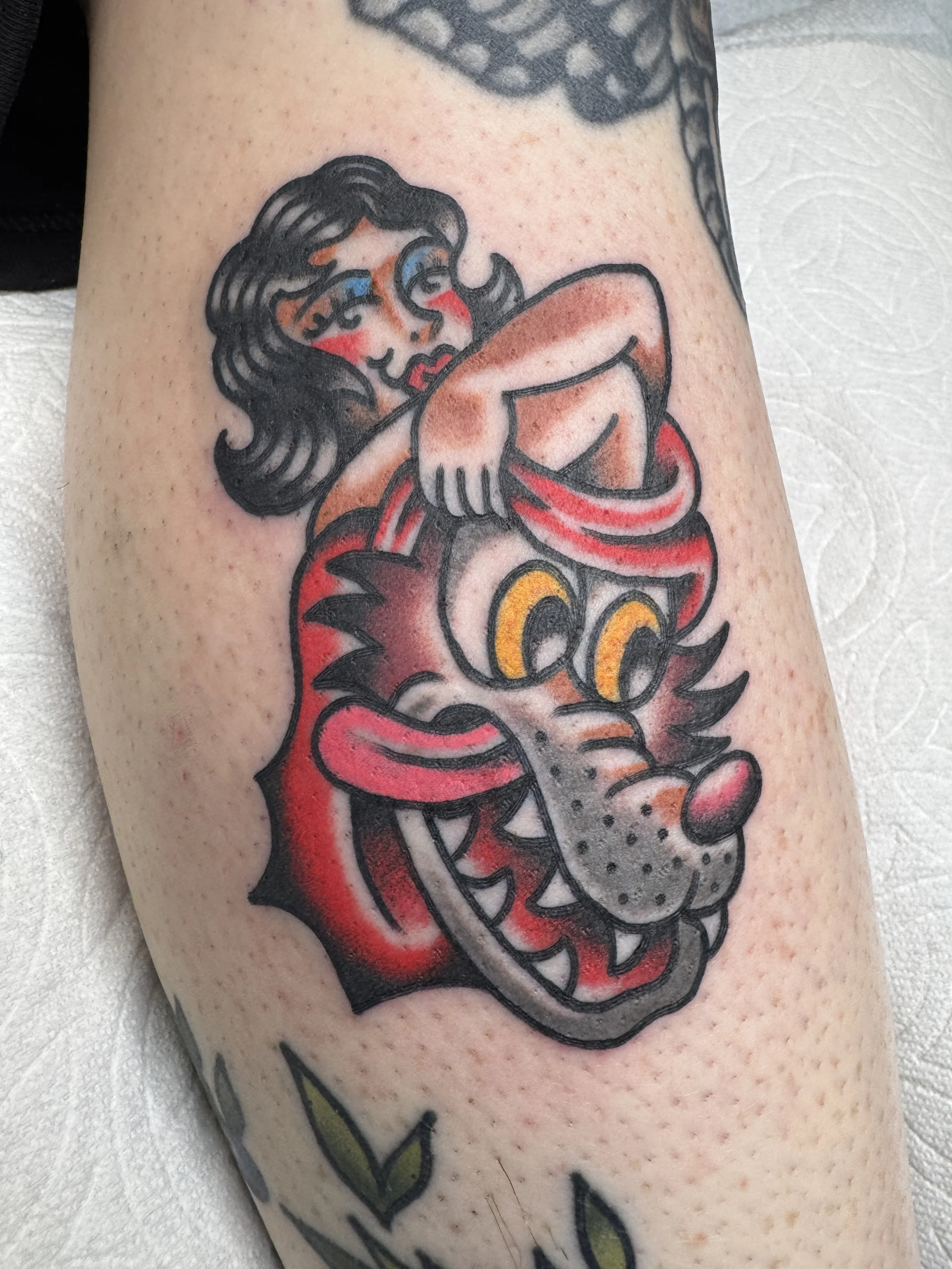 Tattoo of a cartoon woman sitting on a dragon's head with exaggerated features, black outlines, and vibrant colors.