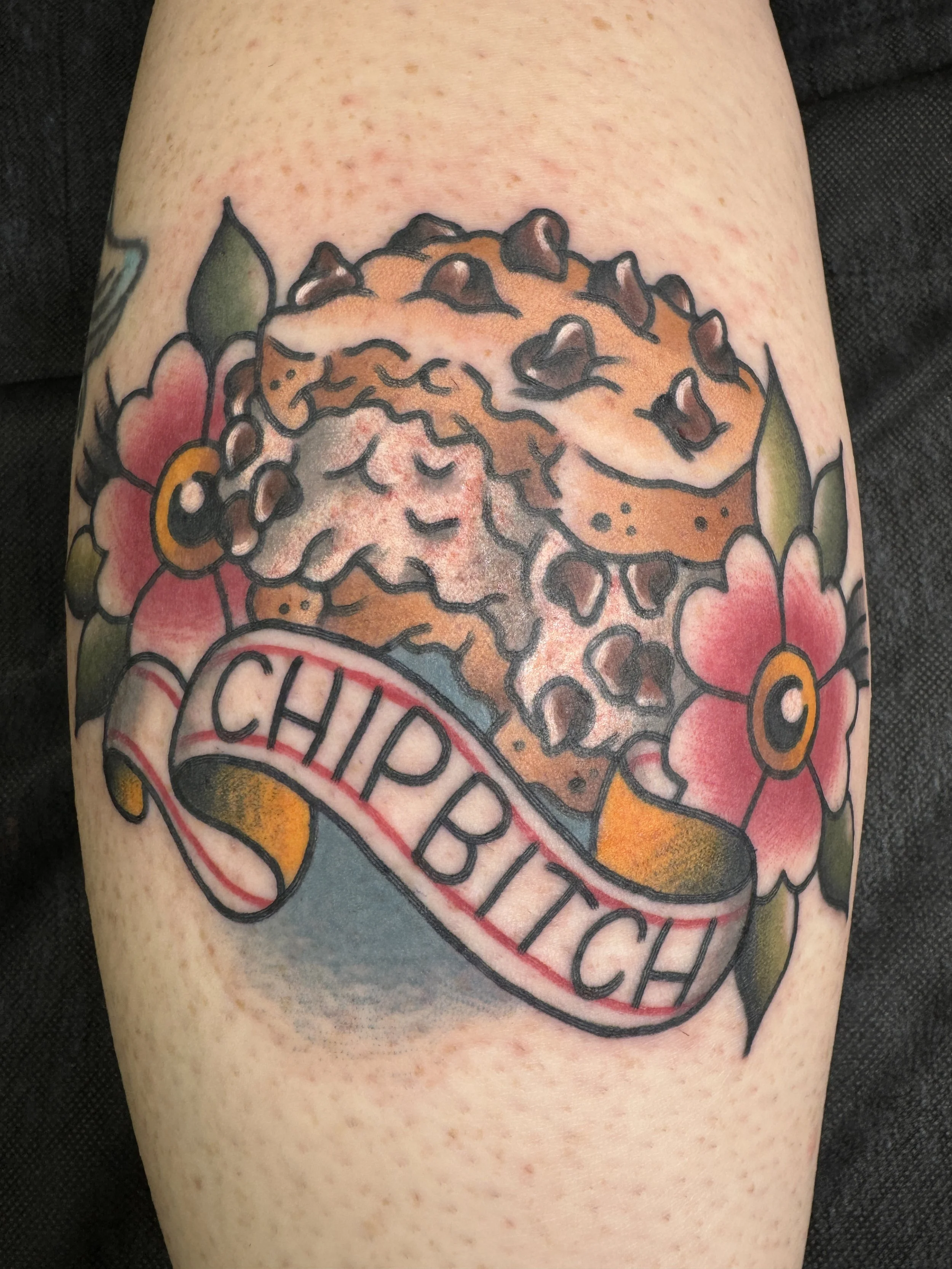 Tattoo of a chocolate chip cookie with pink flowers and green leaves, and the word 'CHIPBITCH' written on a ribbon.
