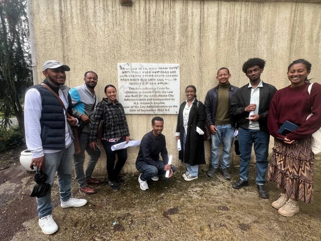 A group of nine people standing outdoors near a beige wall with a plaque, smiling for a photo.