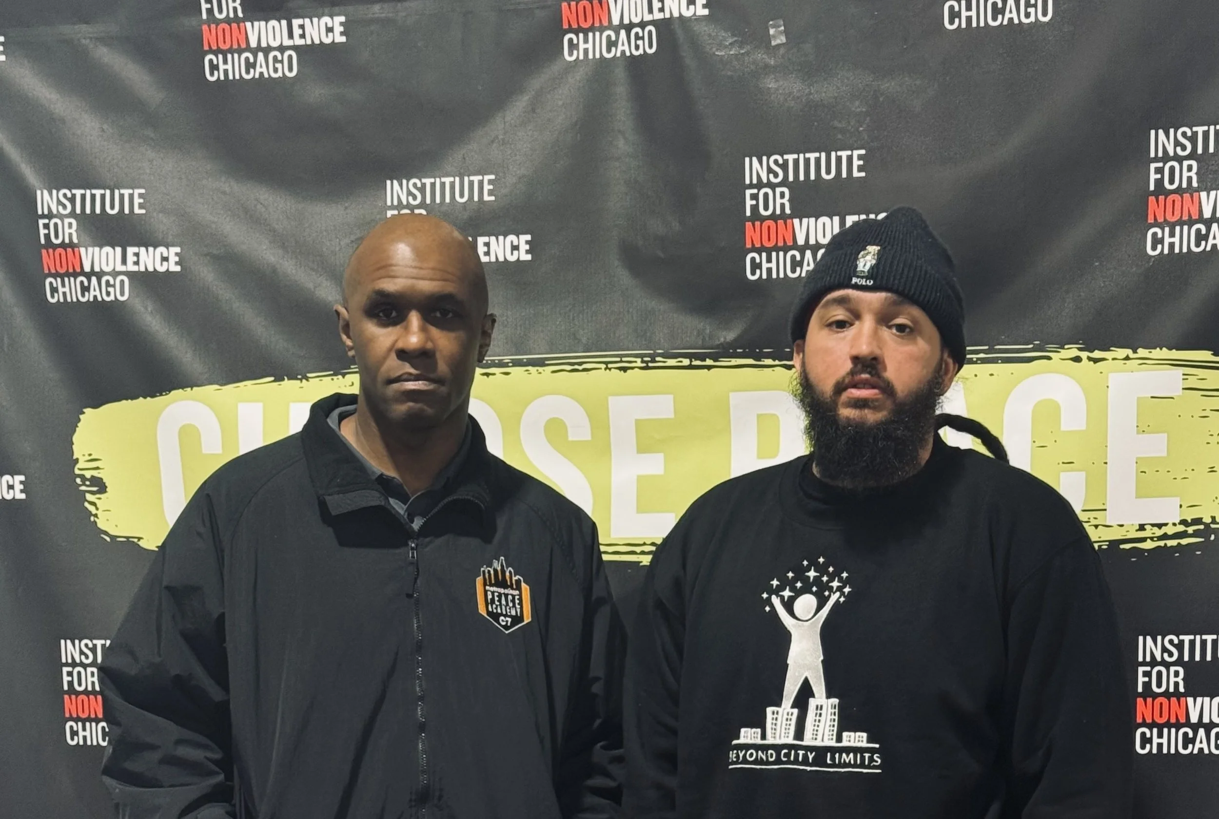 Two men standing in front of a black backdrop with white and red text that reads 'Institute for Nonviolence Chicago.' The man on the left is bald and wearing a black jacket with a logo that says 'Peace City G7' on the left chest. The man on the right has a beard, is wearing a black beanie with a logo, and a black shirt with a white design related to city limits or a skyline.