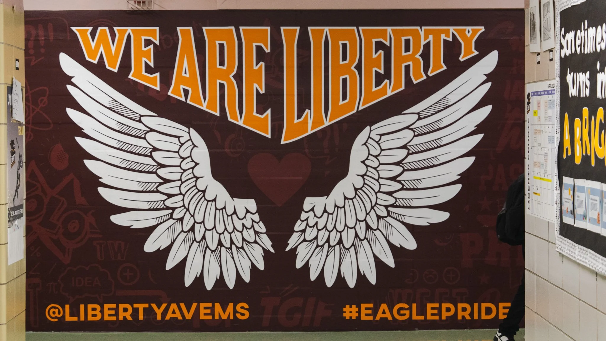 Decorative display with a large white eagle with spread wings, orange and white text that reads "WE ARE EAGLES," and social media handle '@LIBERTYVAVEMS' with the hashtag '#EAGLEPRIDE'.