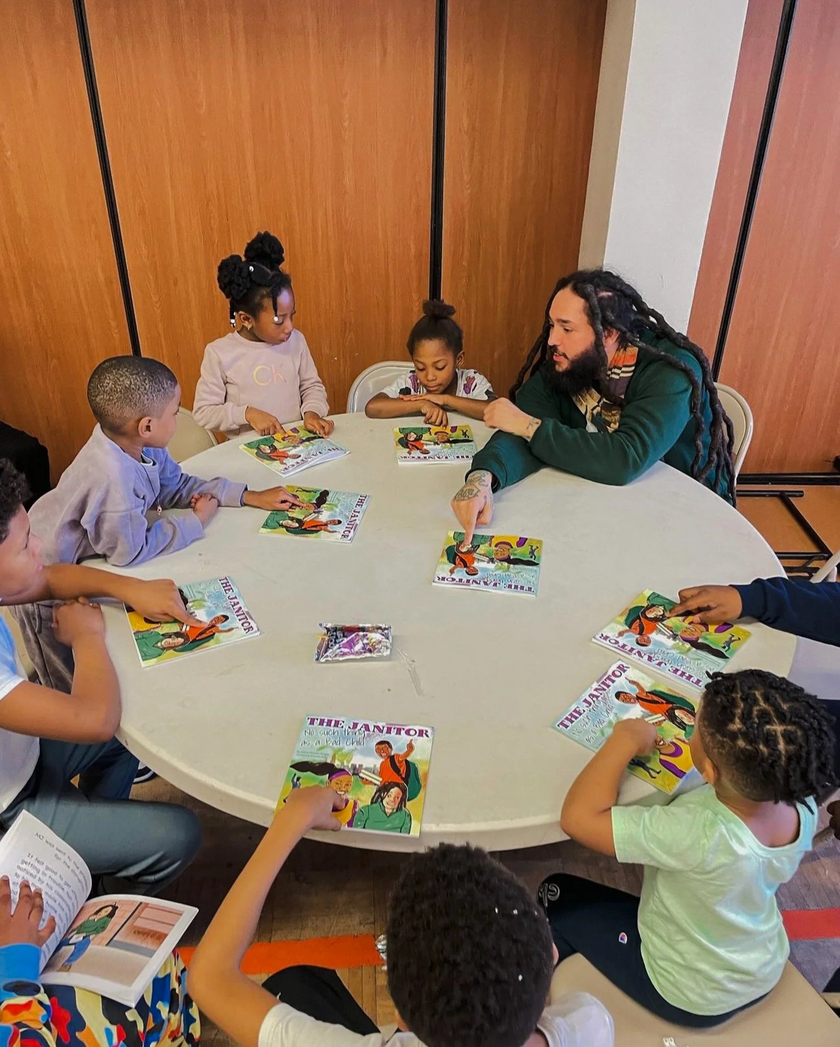 Children sitting around a table reading comic books from 'The Janitor' series, with a man engaging with them, in an indoor setting with wooden panel walls.