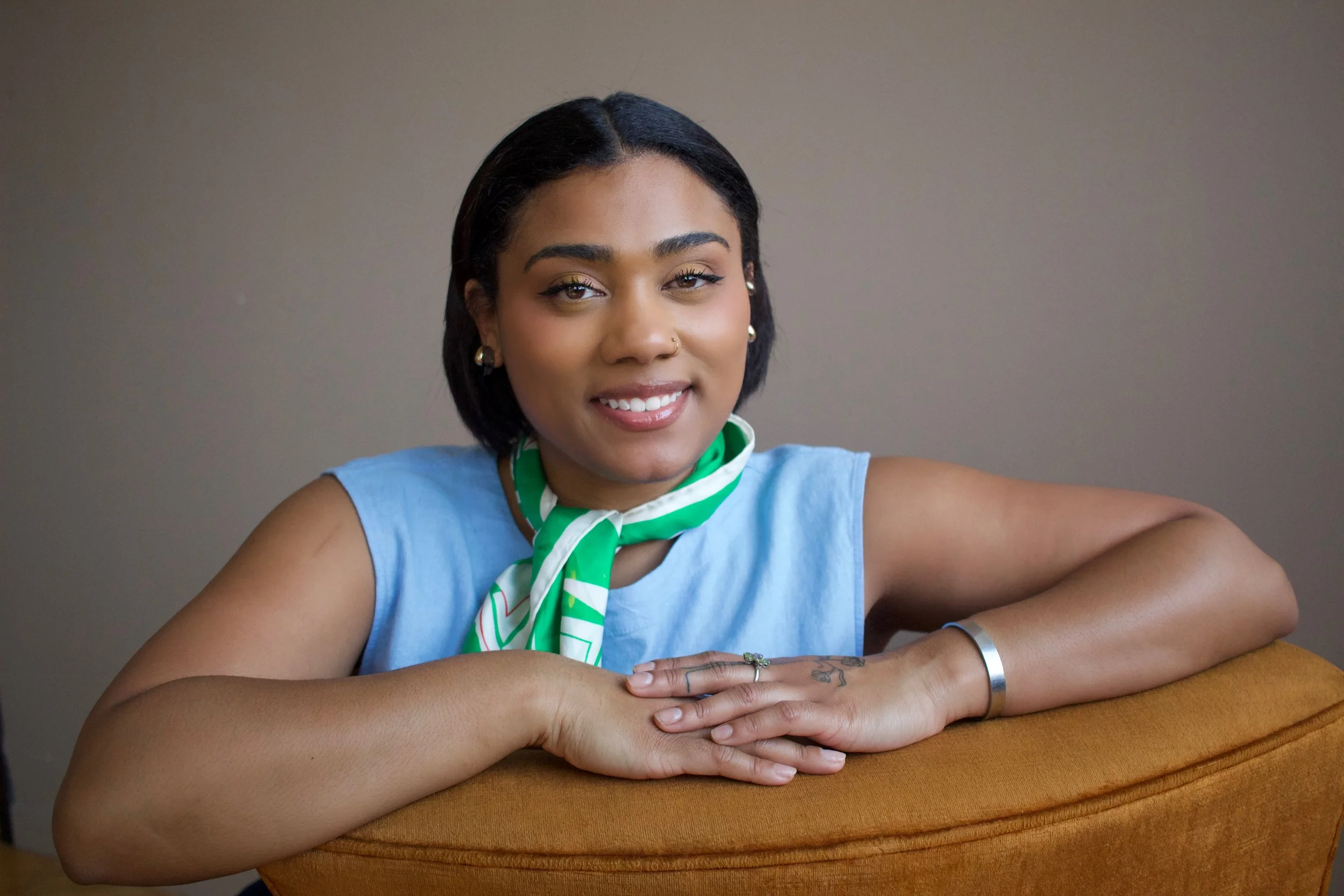A woman with short black hair, wearing a sleeveless light blue top, a green and white scarf around her neck, earrings, and a bracelet, smiling while leaning on an orange cushion.