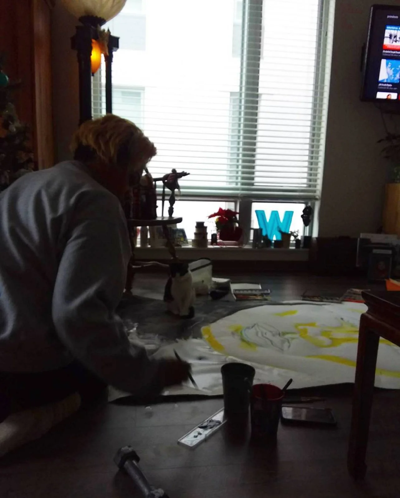 Person painting on a large canvas in a dimly lit room with a window, shelves, and a television.