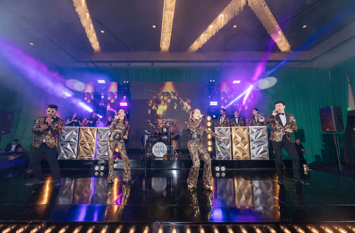 A live band performs on stage at an event, with three singers in metallic jackets singing into microphones and a full band playing behind them. The stage has colorful lights and a large disco ball image on a digital screen.