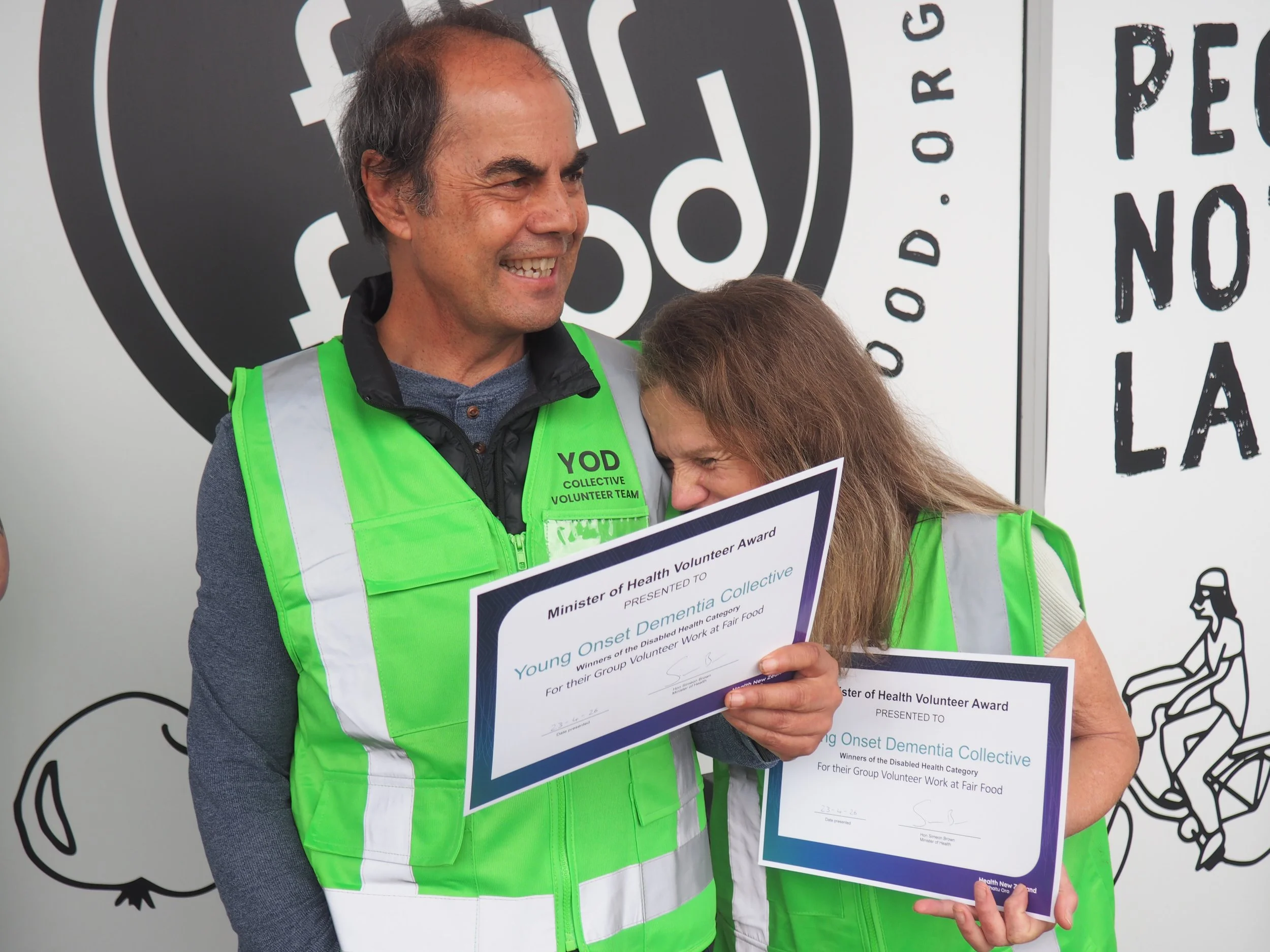 Innes and Anita celebrate their awards outside the Fair Food Hub. They are in green high vis and holding an award certificate while smiling.