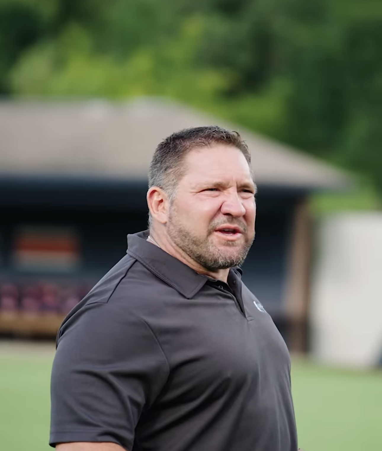 A man with short dark hair and a beard looking to the side outdoors, wearing a black polo shirt.