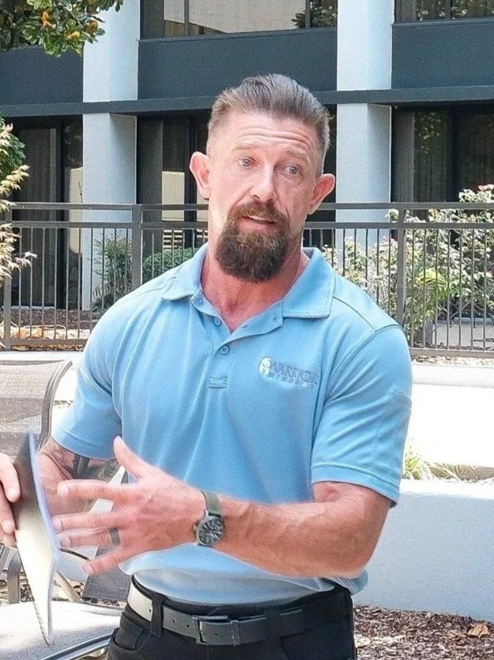 A man wearing a light blue polo shirt with a logo, standing outdoors in front of a modern building with windows, a fence, and some greenery.