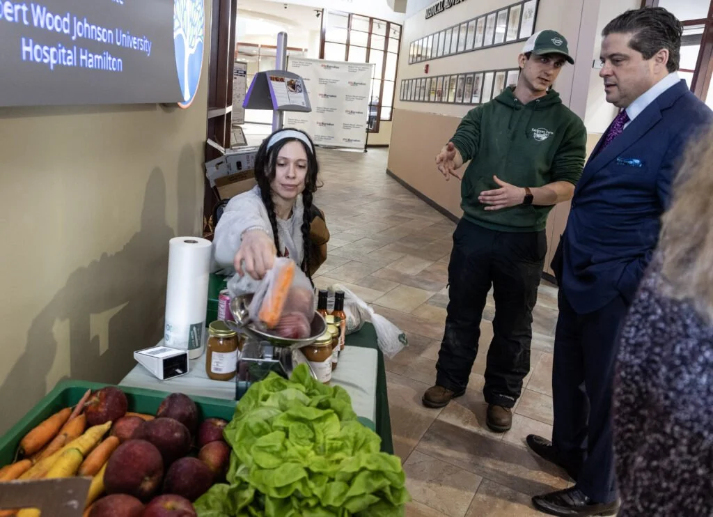 A woman demonstrating fresh produce at a health fair, with a display of apples, carrots, lettuce, and jars of preserves, while three men, including one in a suit, observe and listen in a hospital corridor.