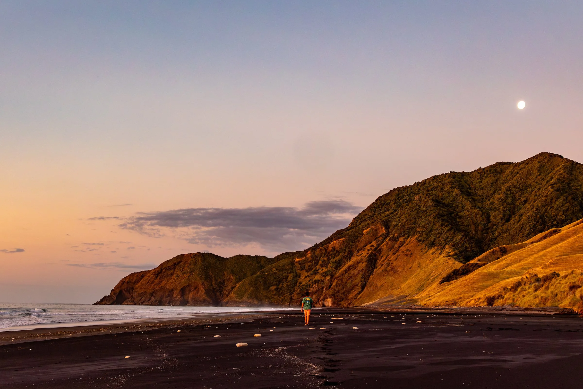 Scenic view of a grassy coastal area with a dark structure and a wooden deck, overlooking the ocean at sunset. The sky is cloudy with vibrant colors on the horizon.