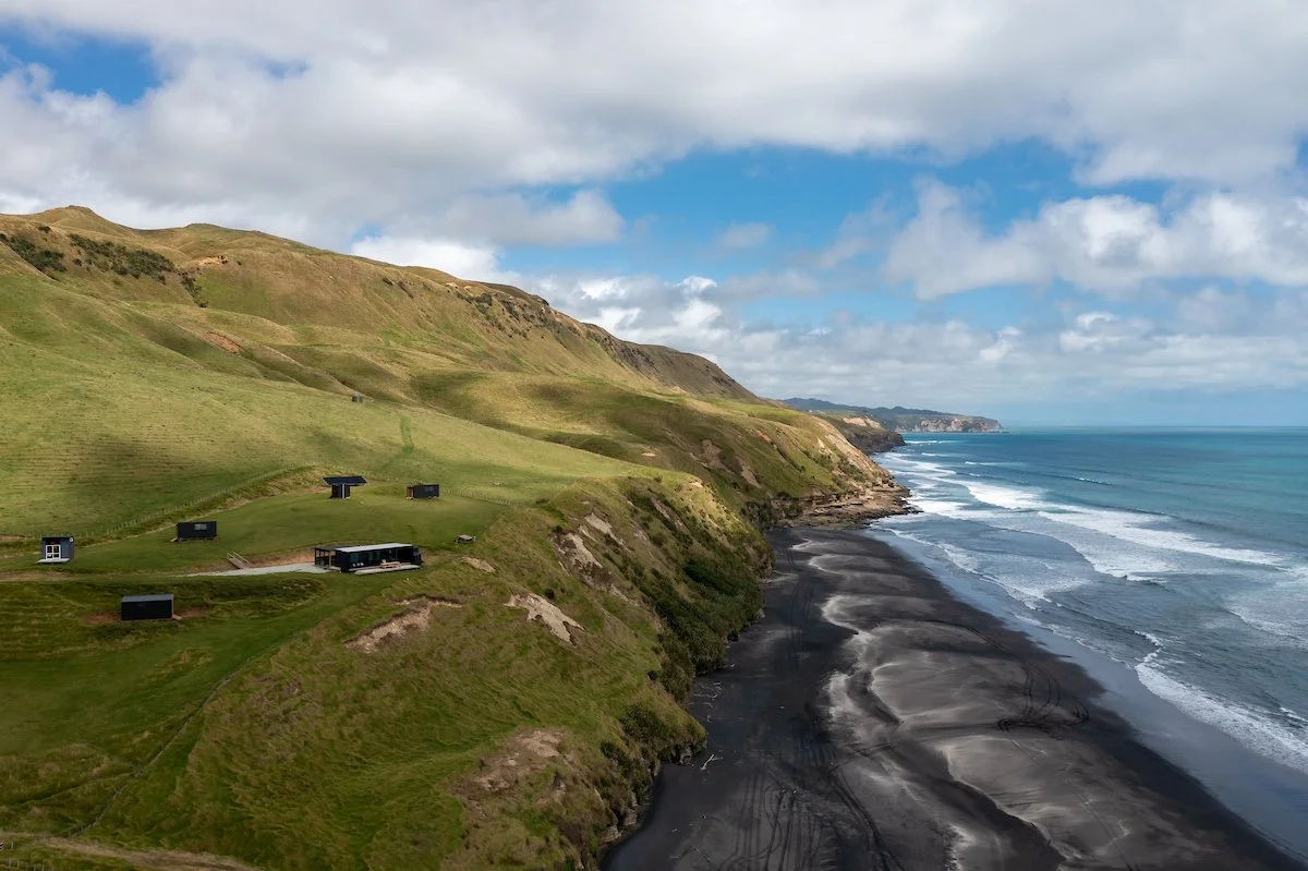 Aerial view of a black sand beach bordered by green rolling hills and the ocean.