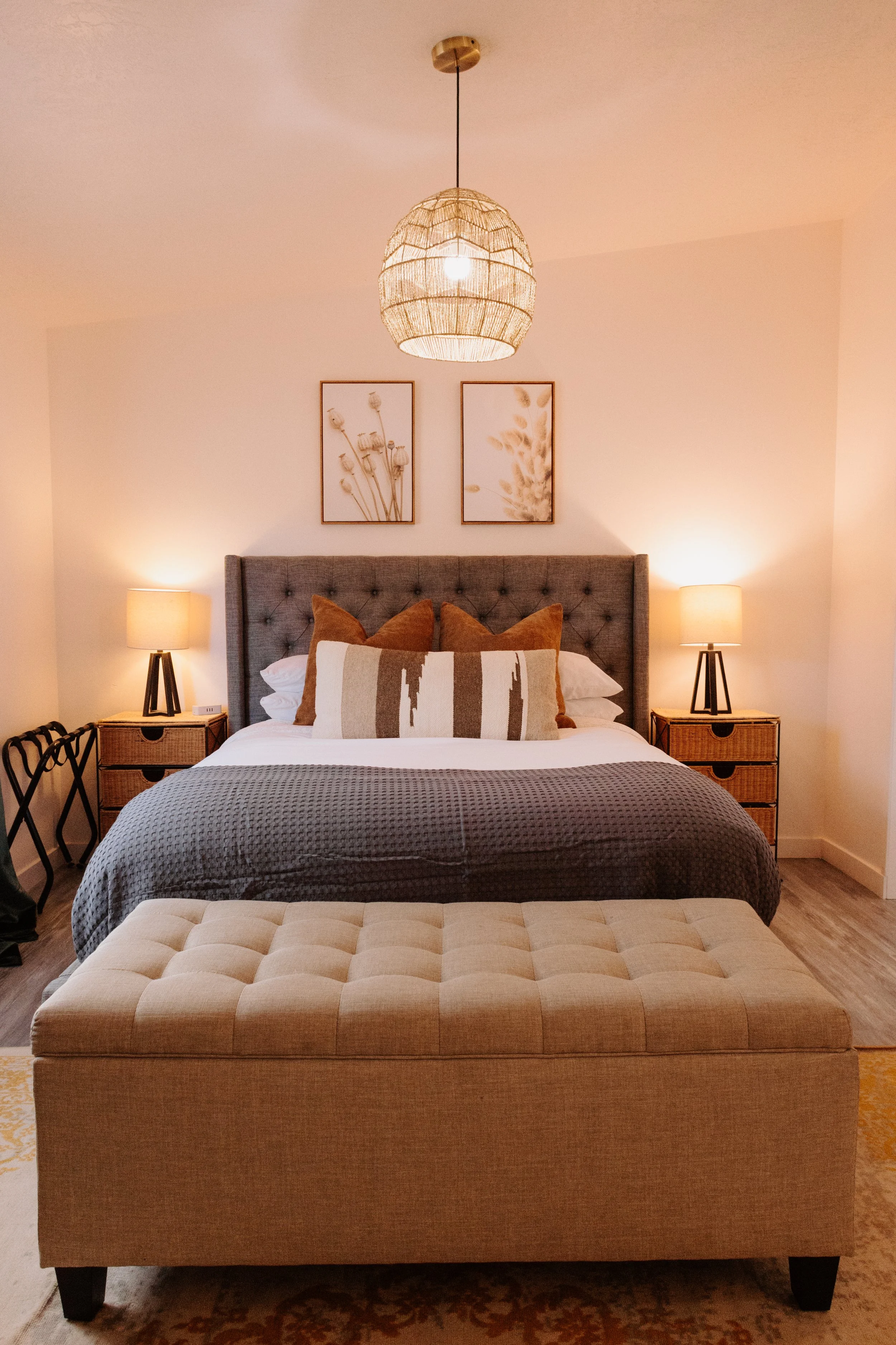 A neatly made bedroom with a large bed with a tufted gray headboard, decorated with brown and beige pillows. There are two wooden nightstands with lamps on each side of the bed. Above the headboard, two framed botanical prints are hung. At the foot of the bed, a beige tufted bench sits on a patterned rug, and a woven pendant light hangs from the ceiling.