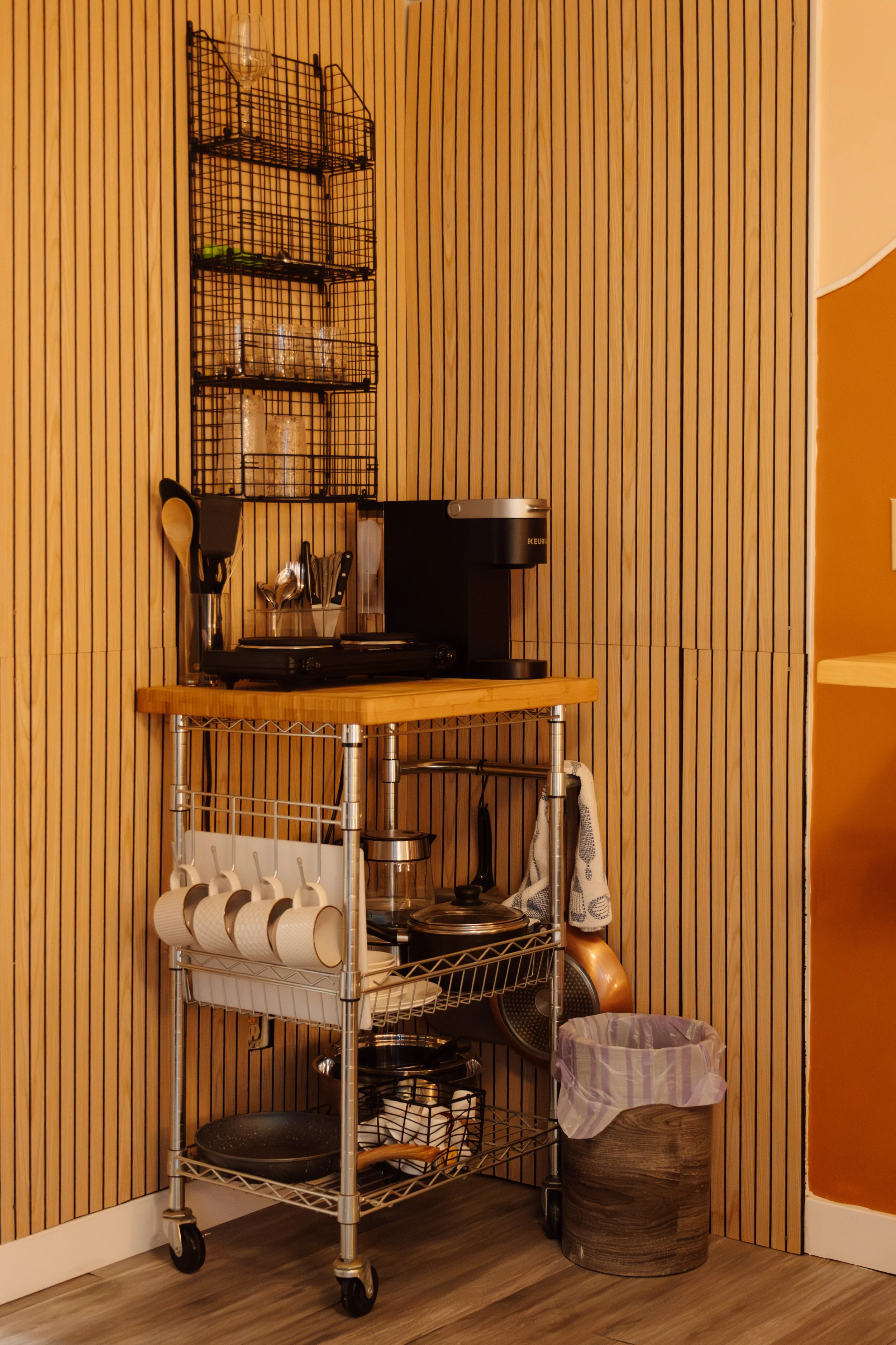 A kitchen corner with wooden wall paneling, a metal rolling cart holding cups, pots, and kitchen utensils, a small wooden table with a coffee maker, and a wall-mounted wire shelf with glasses and jars.