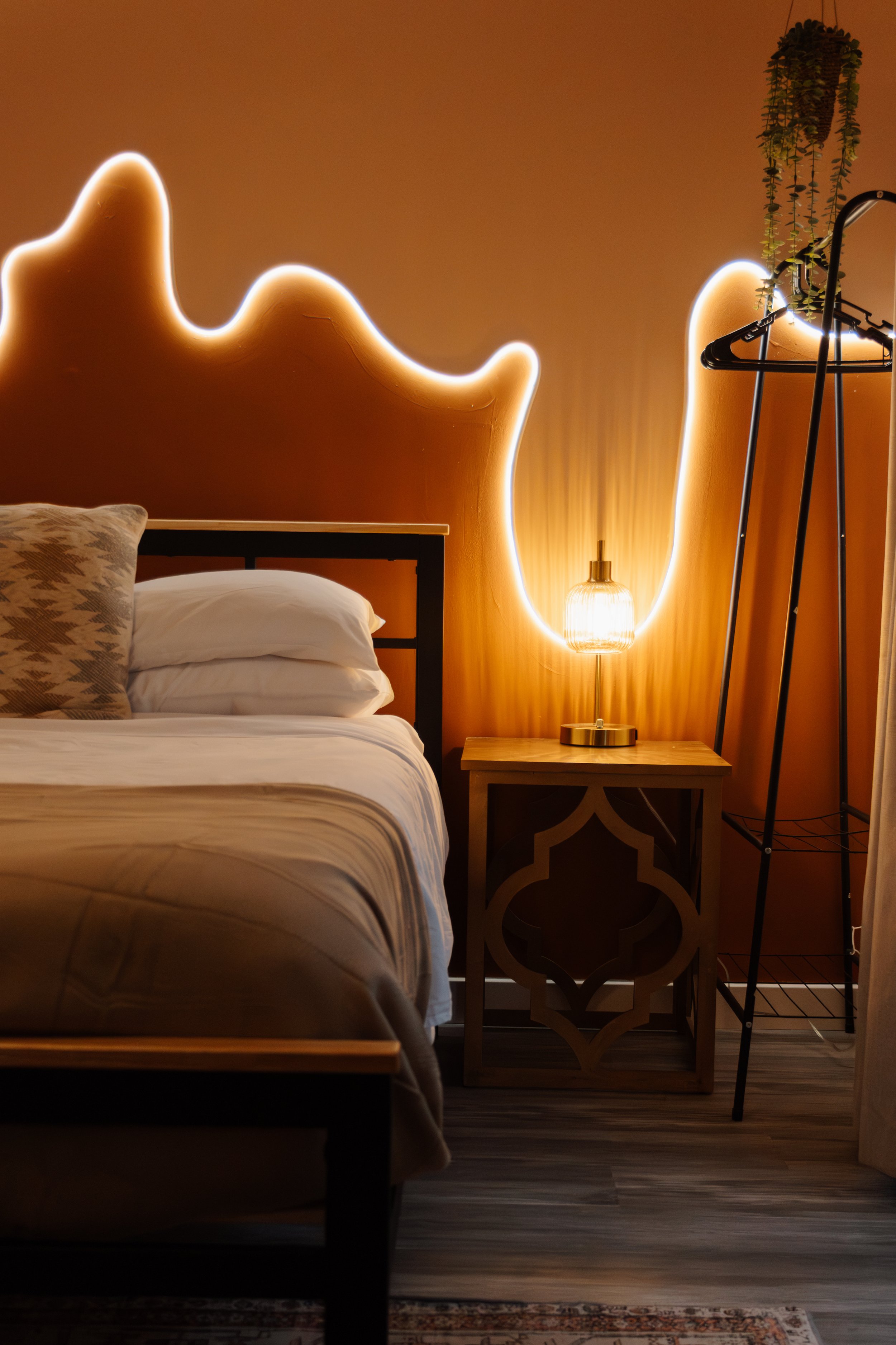 A bedroom with a bed, a nightstand and a lamp. The bed has white bedding and a patterned pillow. The headboard has an abstract, glowing, neon-like outline. A plant hangs from the ceiling, and an open clothing rack stands nearby.