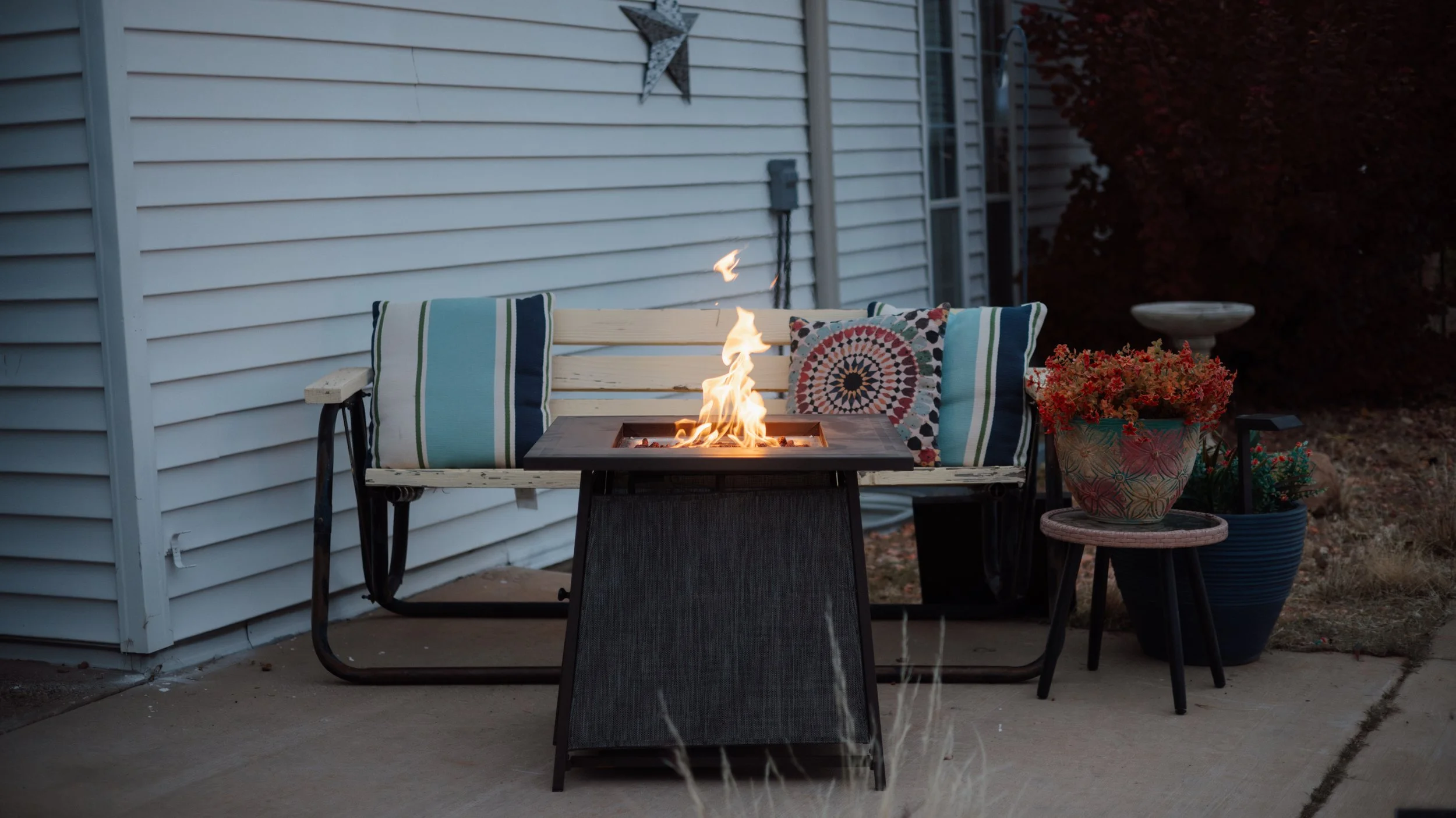 Outdoor patio scene with a bench, colorful pillows, a fire pit, and potted plants beside a house with white siding.