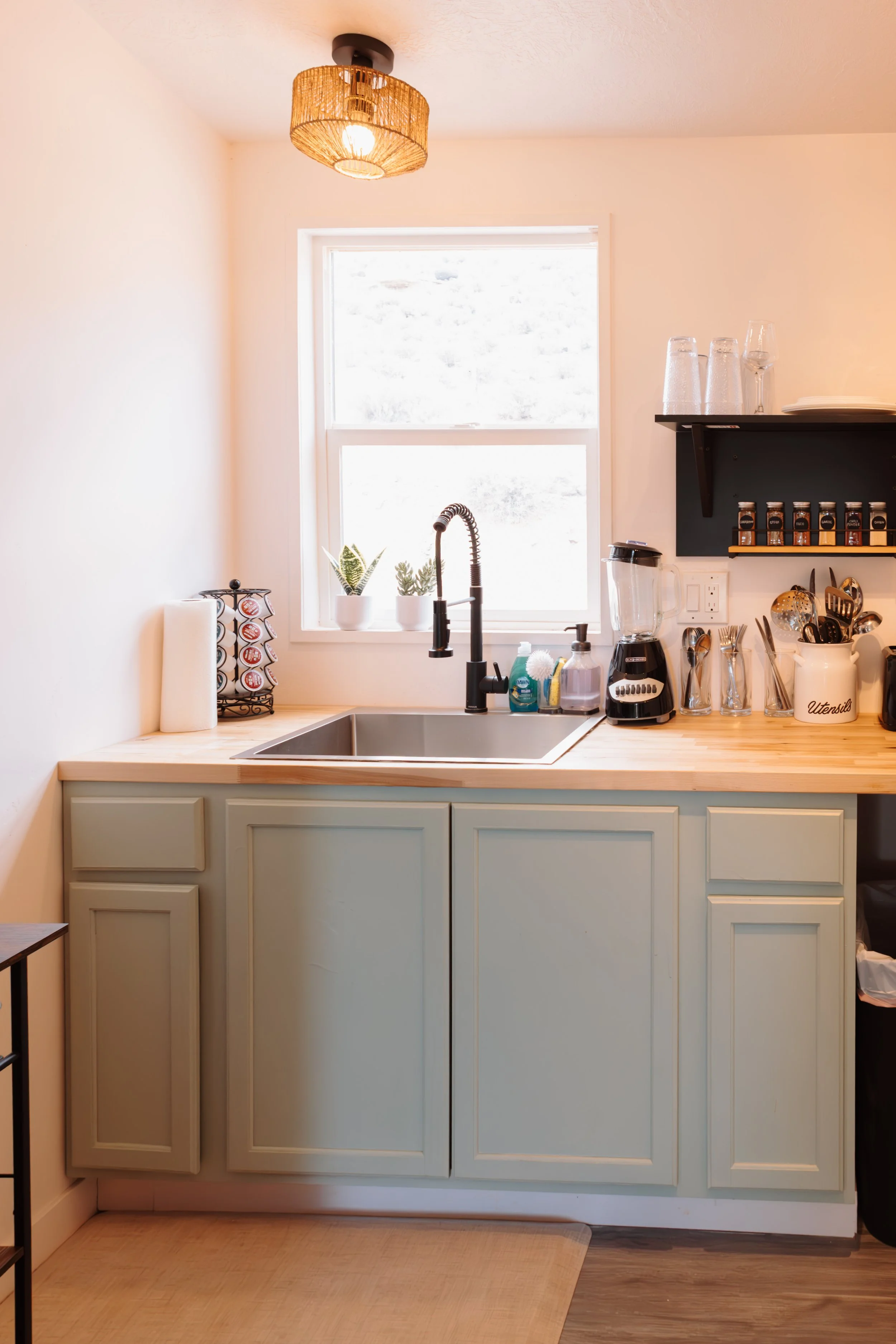 A minimalist kitchen with a single window, light green cabinets, a wooden countertop, a black faucet, and various small appliances and utensils on the countertop. There's a light fixture on the ceiling.