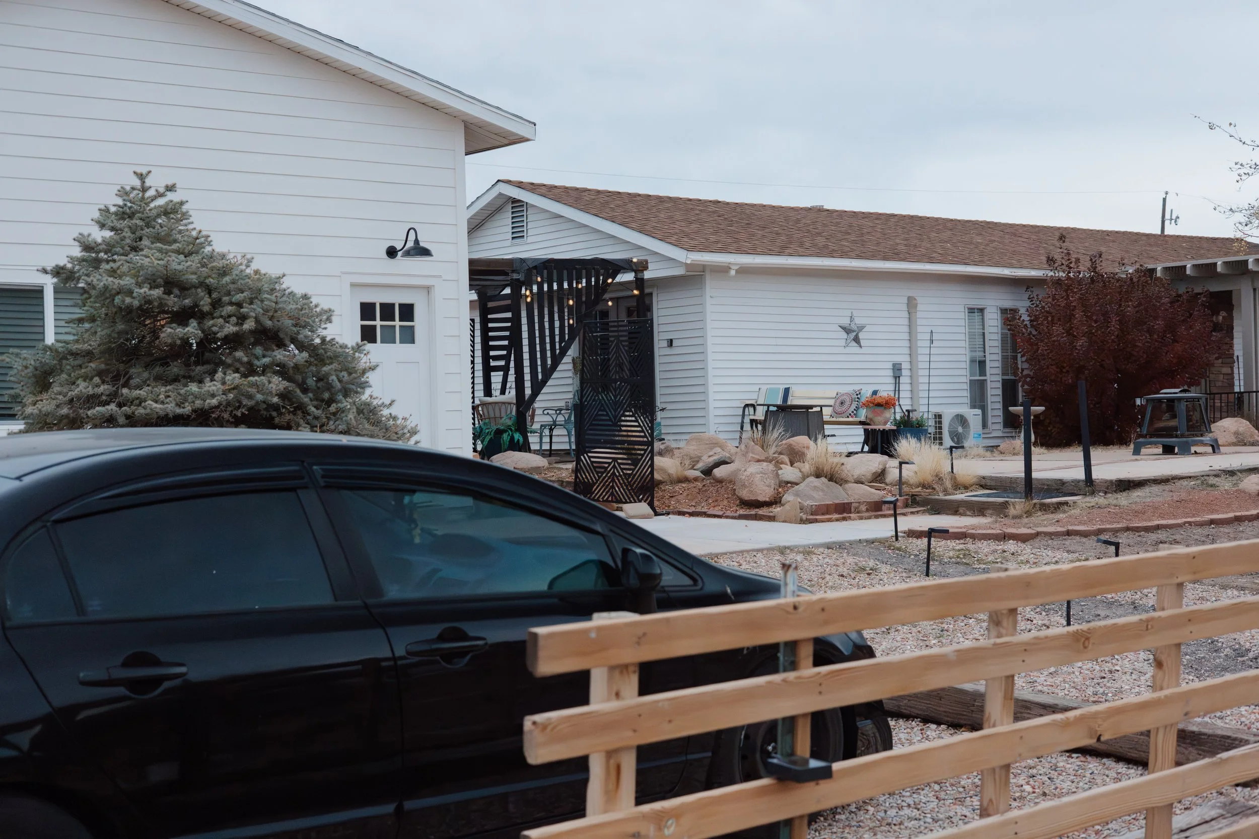 Corner of a white house with a small porch, black metal fencing, and outdoor seating, next to a large tree with reddish leaves, and a black car parked in the foreground.