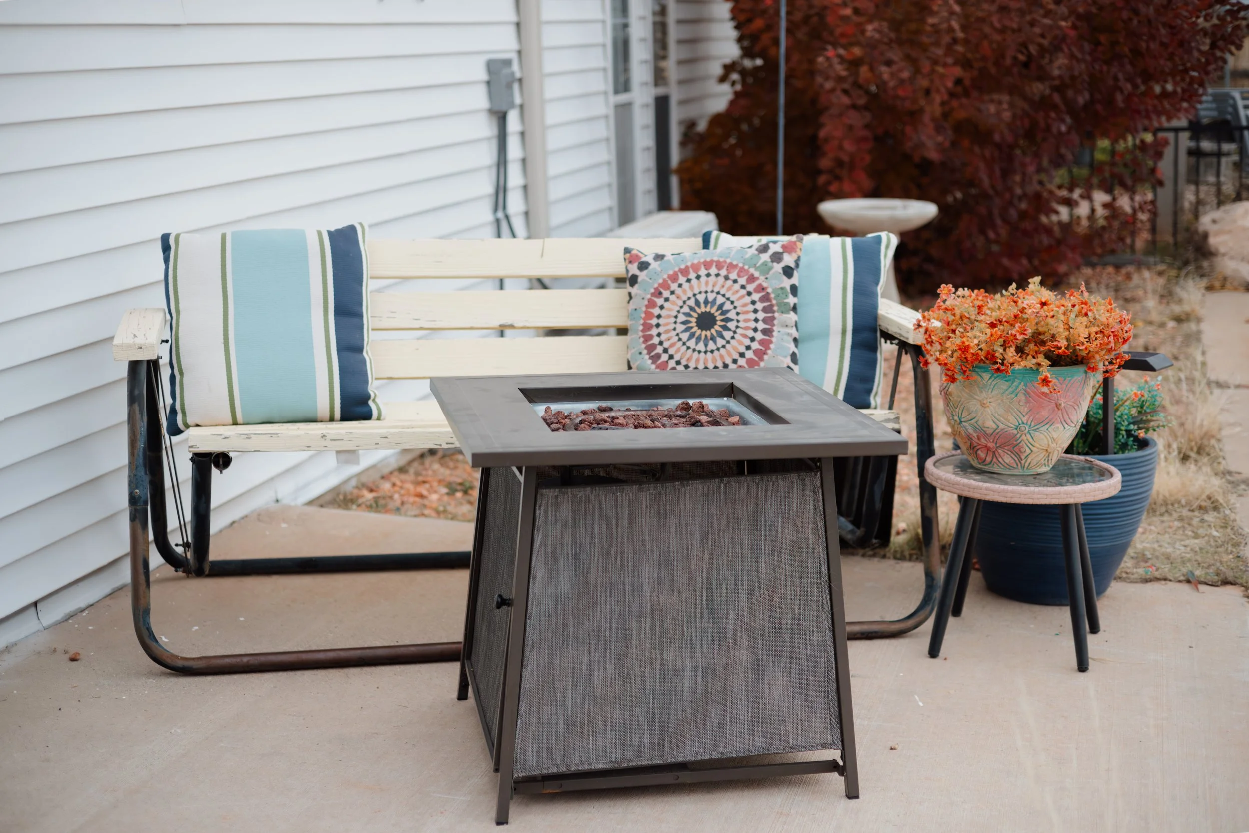 A patio scene with a white wooden bench with striped throw pillows, a black firepit table, and potted plants with orange flowers, against a house with red autumn leaves in the background.