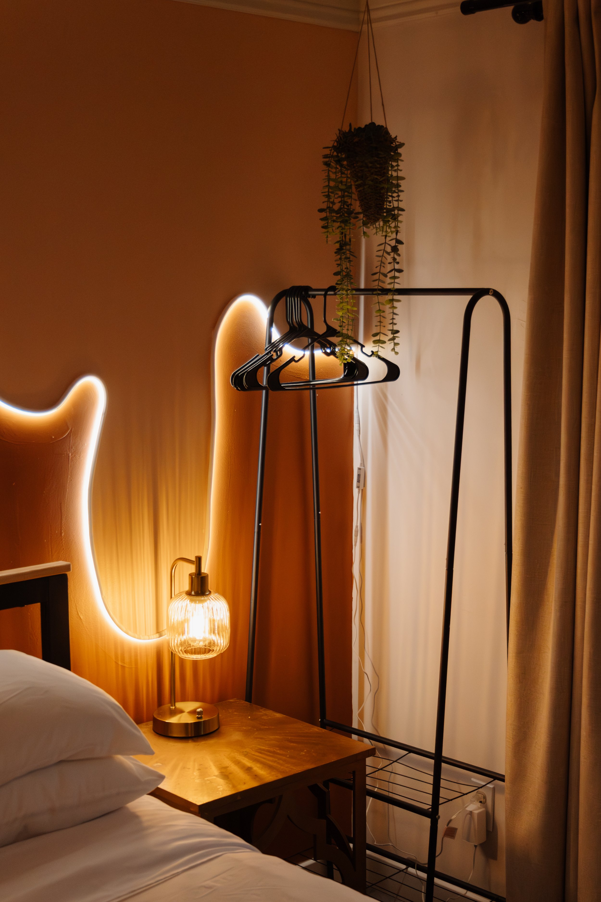 A bedroom corner with a bed, a wooden nightstand, a decorative lamp, hanging plants, and a black clothing rack against an orange wall with a neon light accent.