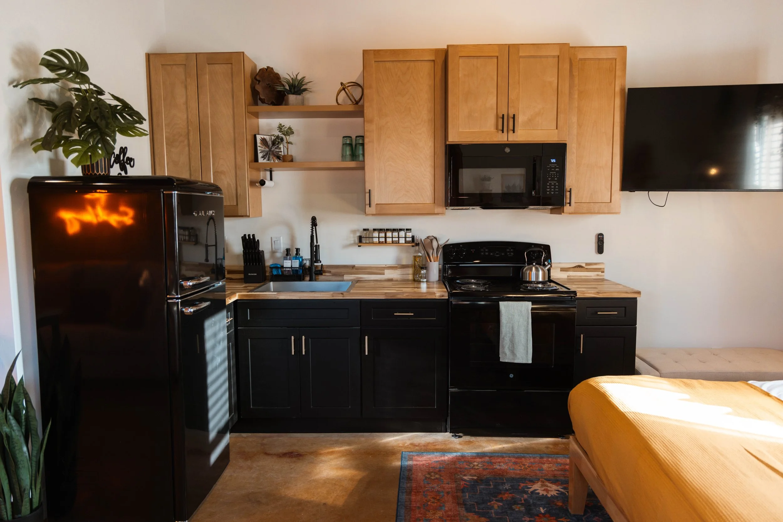 A compact kitchen with black lower cabinets, light wooden upper cabinets, and a wooden countertop. There is a black refrigerator, a black stove with a tea kettle, a microwave, and a wall-mounted TV. There are open shelves with decorative items and green plants, and a small window with blinds partially visible.