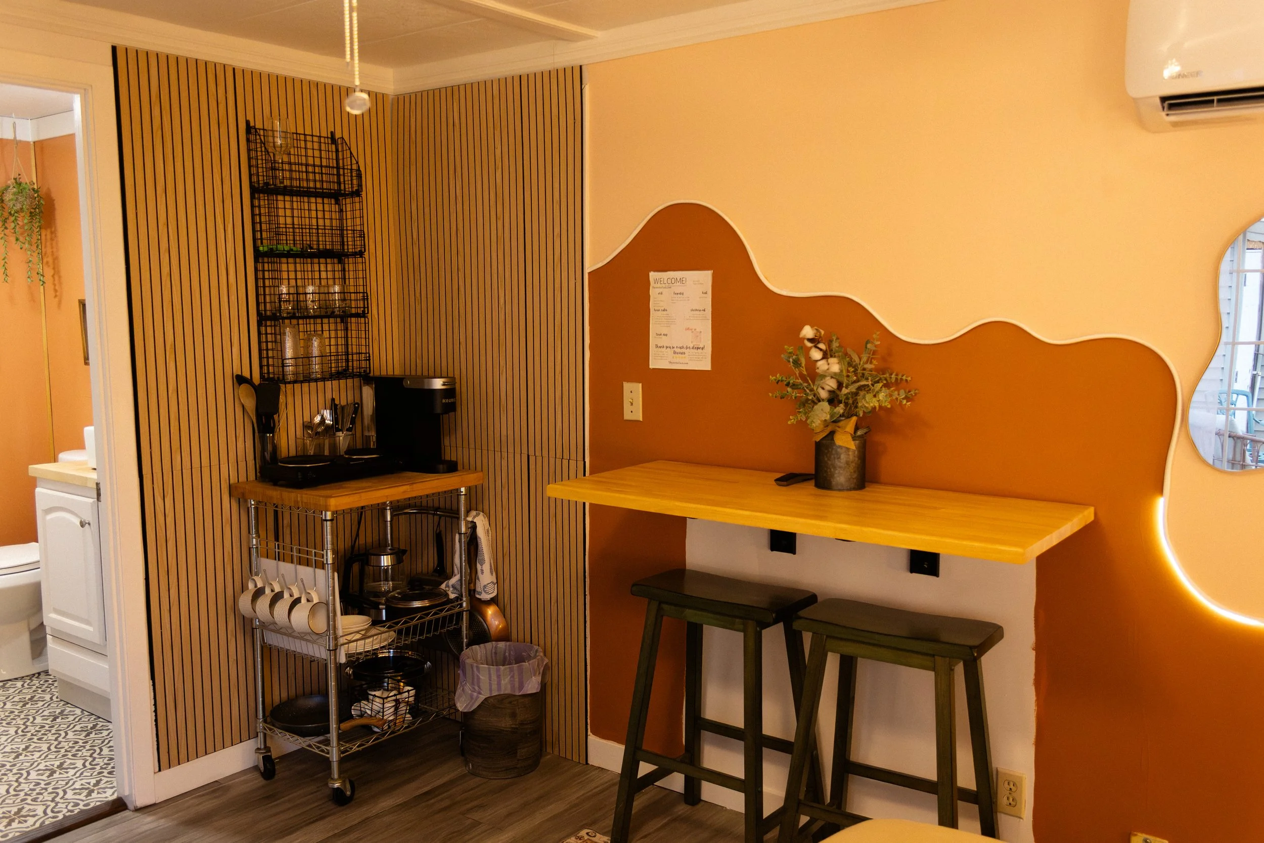 A cozy coffee corner in a cafe with a wooden bar-height table, two dark green stools, a decorative flower arrangement, a wall with a geometric design, and a small shelf with coffee supplies