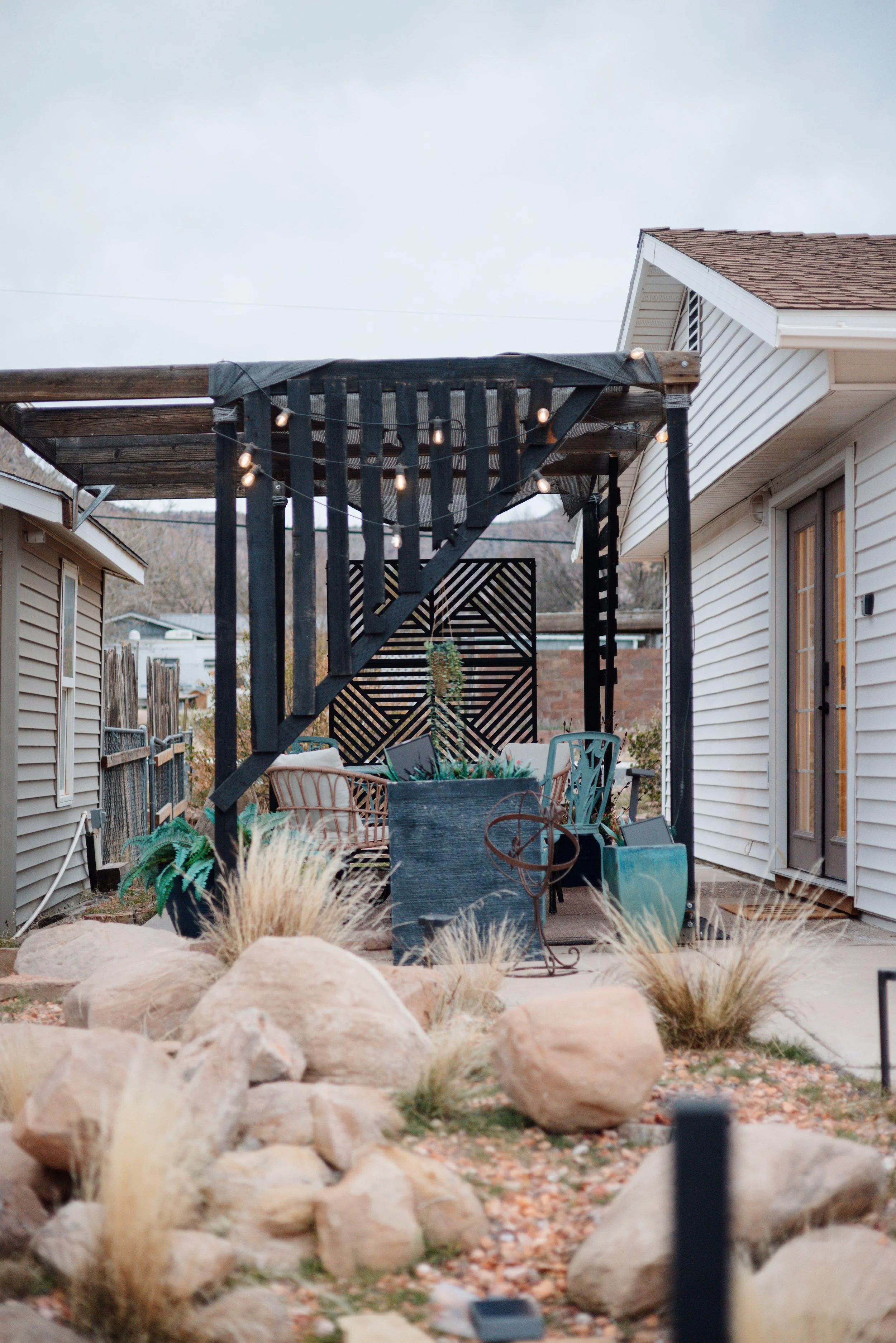 A backyard patio with a wooden pergola, string lights, outdoor furniture, decorative rocks, and dry grass, with a house on the right side and a fence in the background.