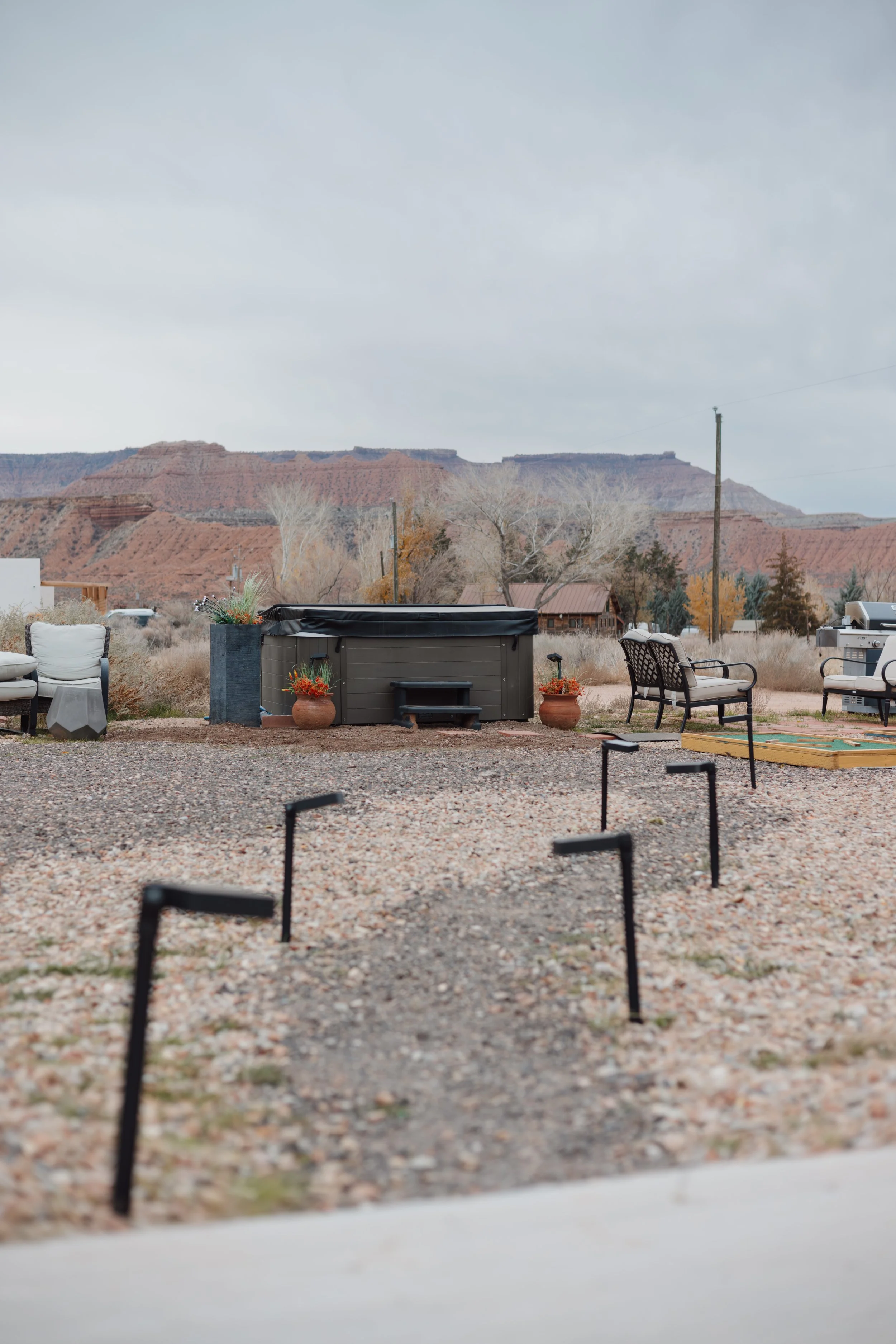 A backyard patio with a hot tub, outdoor furniture, potted plants, a grill, and a gravel ground, with a mountainous landscape in the background.